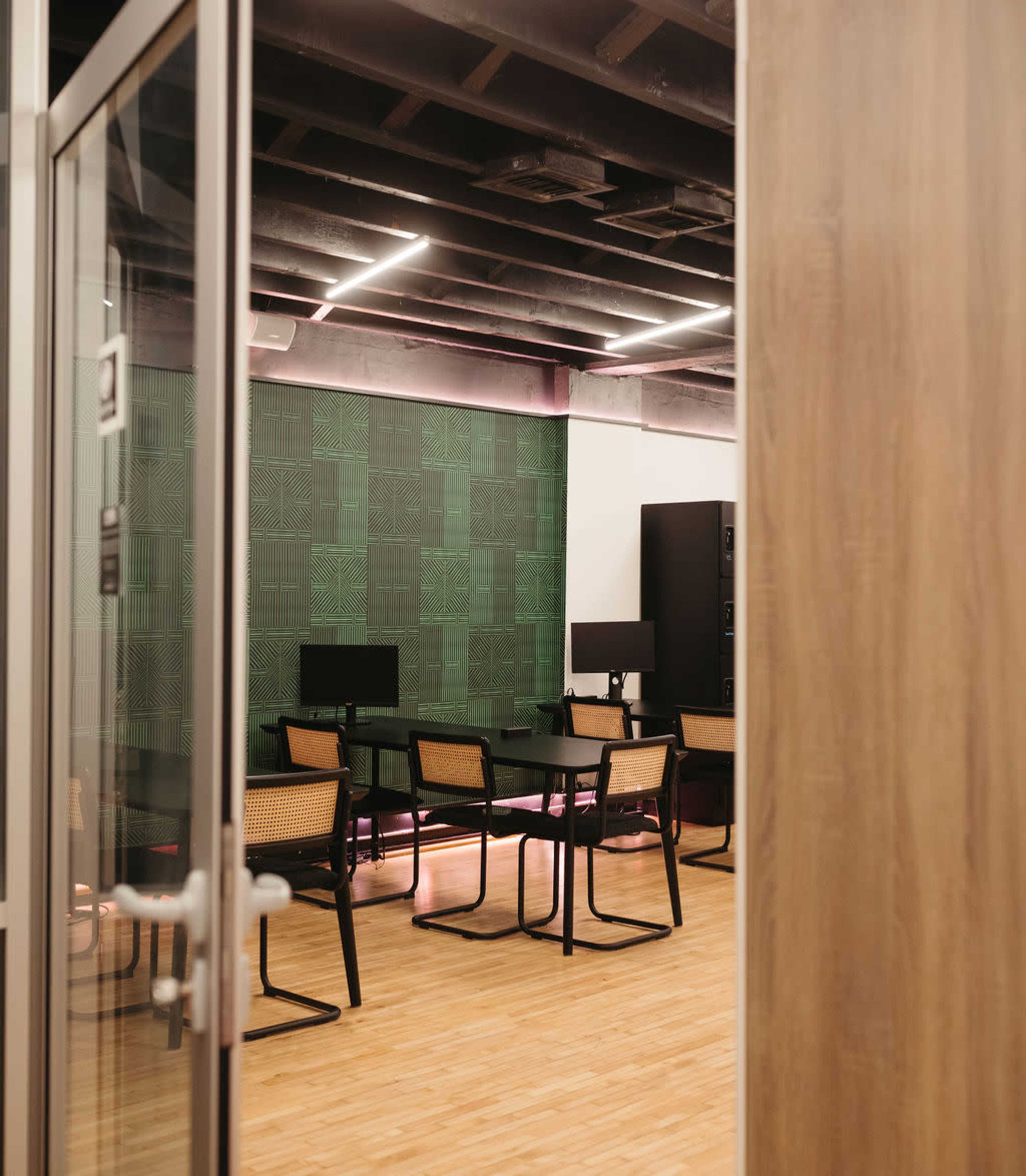 The image shows a modern conference room with a green patterned wall, wooden floors, and multiple black chairs around a long table.