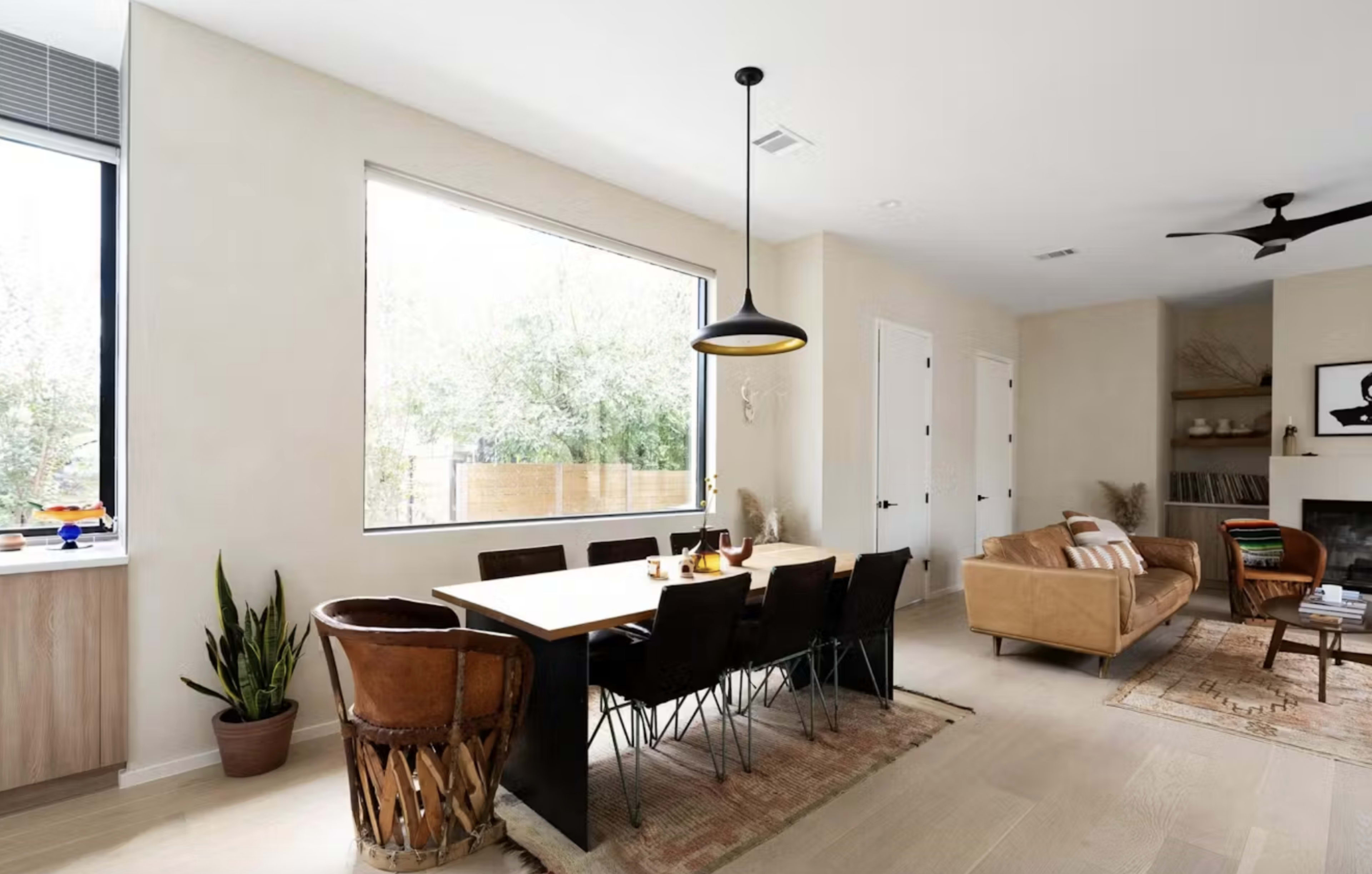 A modern dining area features a long table with a pendant light, surrounded by wooden chairs and a decorative plant, adjacent to a living space with a sofa and a fireplace.
