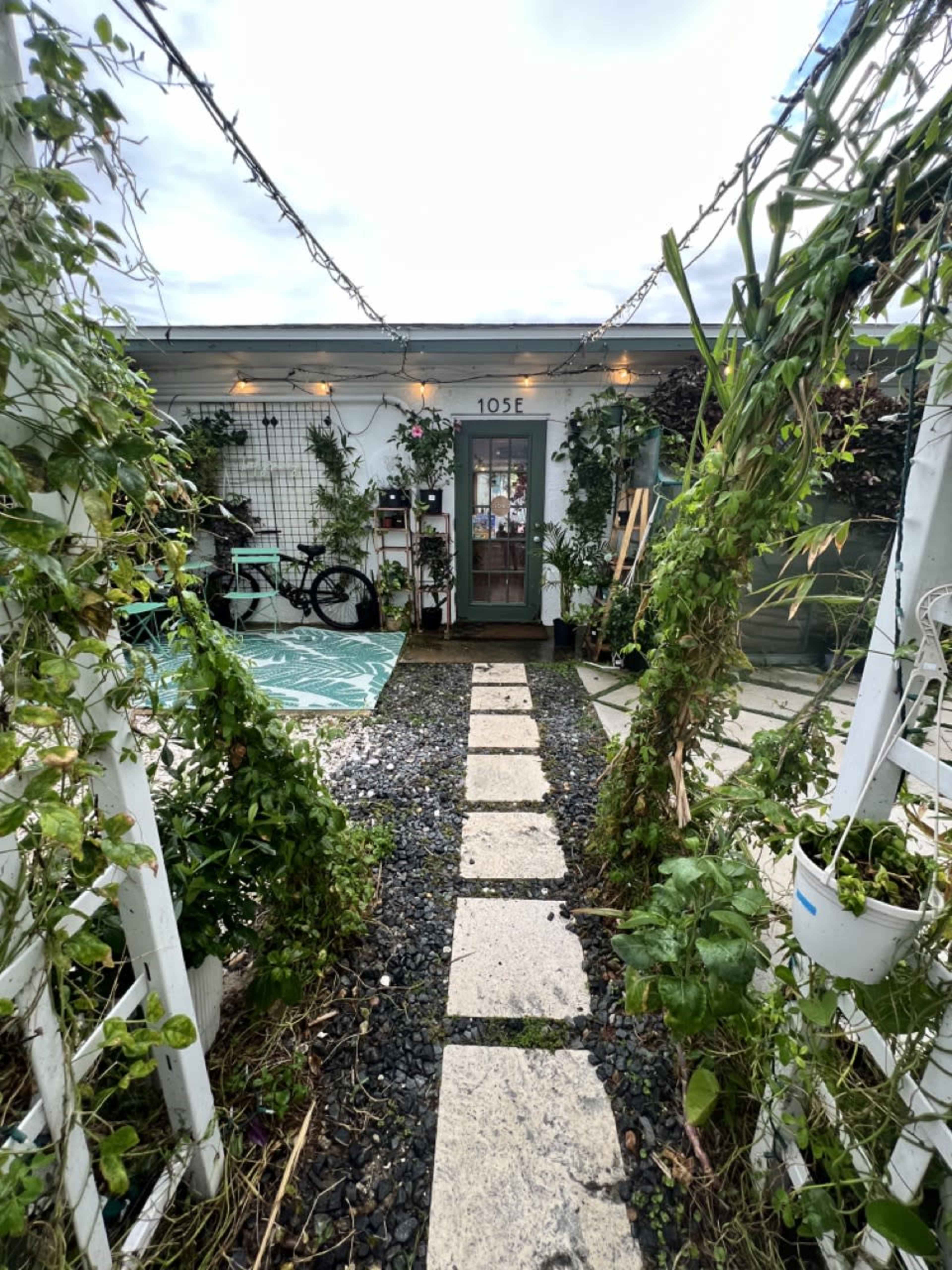 A stone pathway lined with greenery leads to a door surrounded by plants and decorative lights.