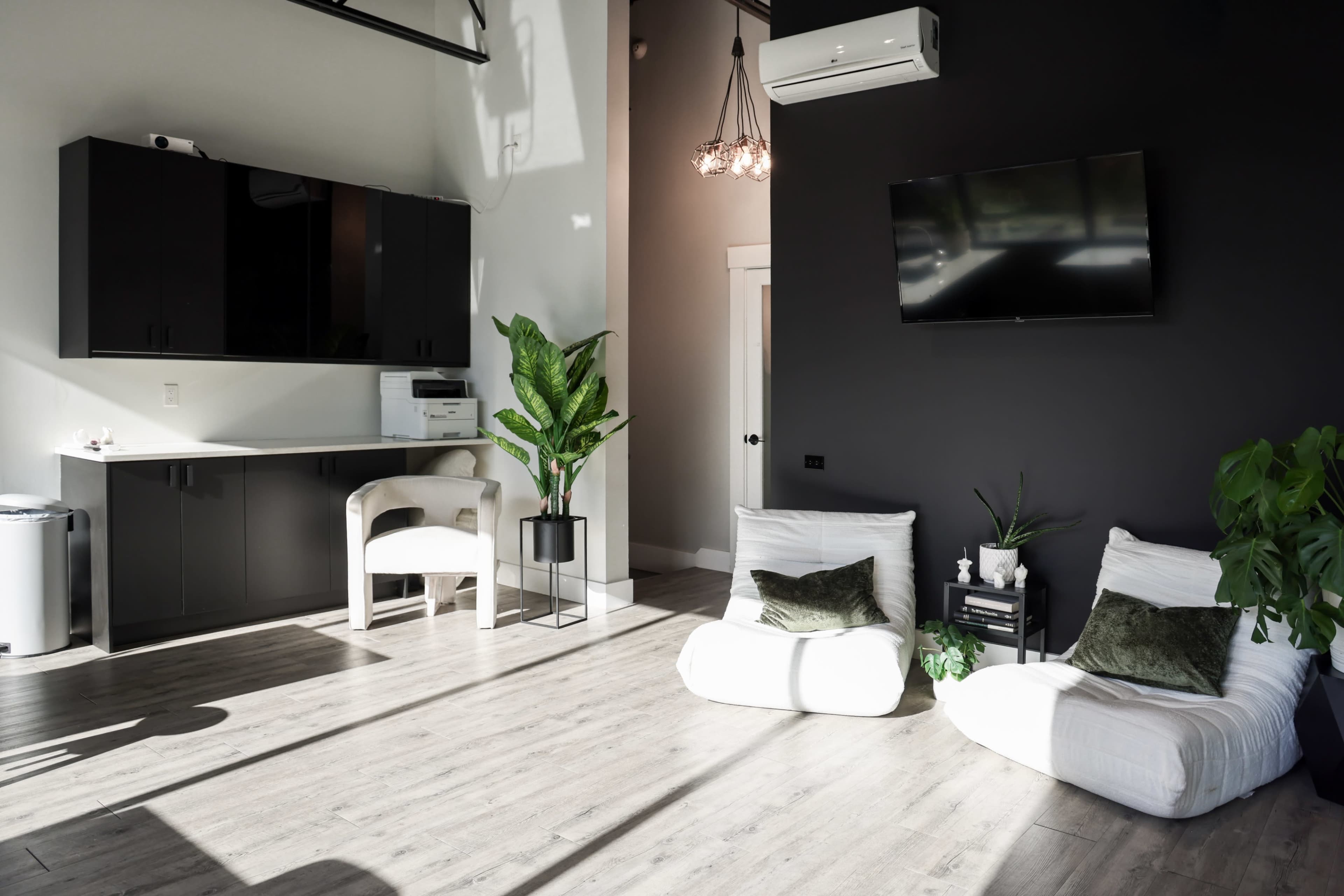 The image shows a modern, minimalistic interior space featuring a wall-mounted TV, two white lounge chairs, a small table with plants, and a black accent wall.