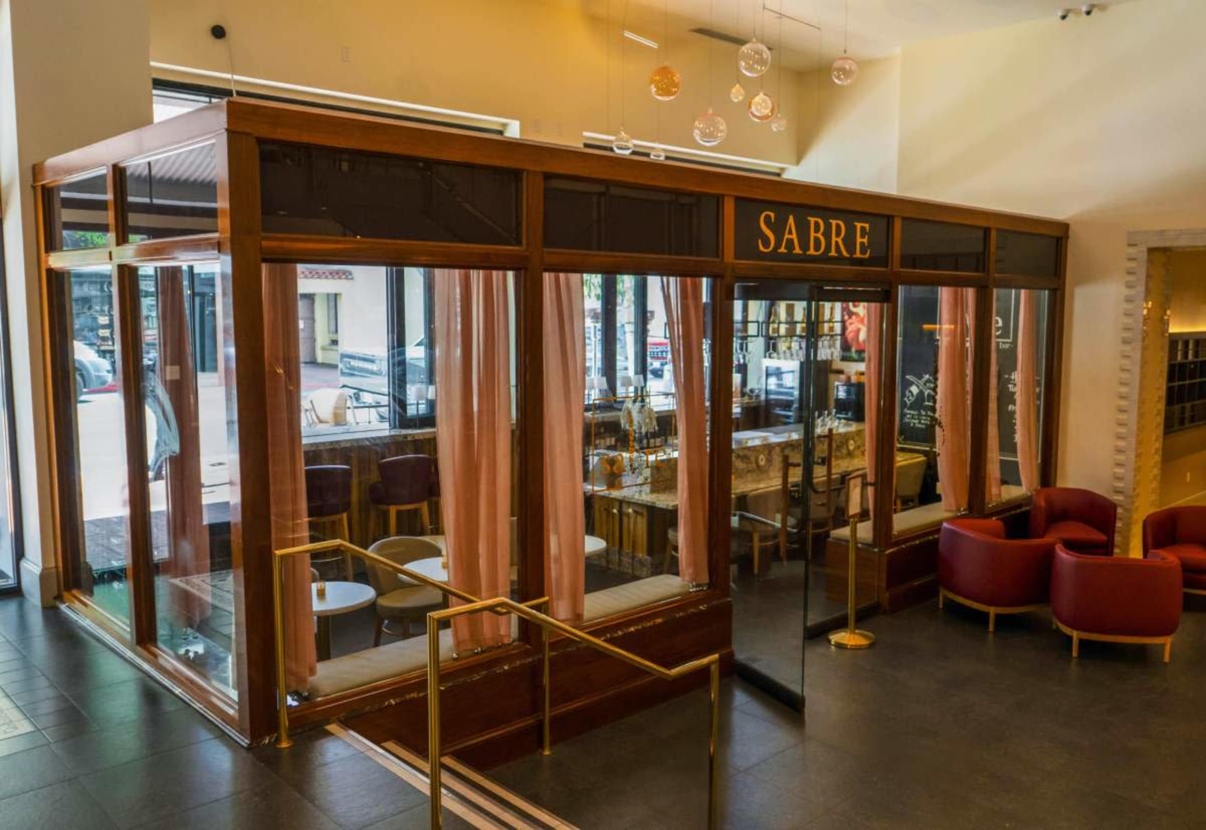 The image shows a modern café or bar with large glass windows, a wooden structure, and neatly arranged seating areas inside.