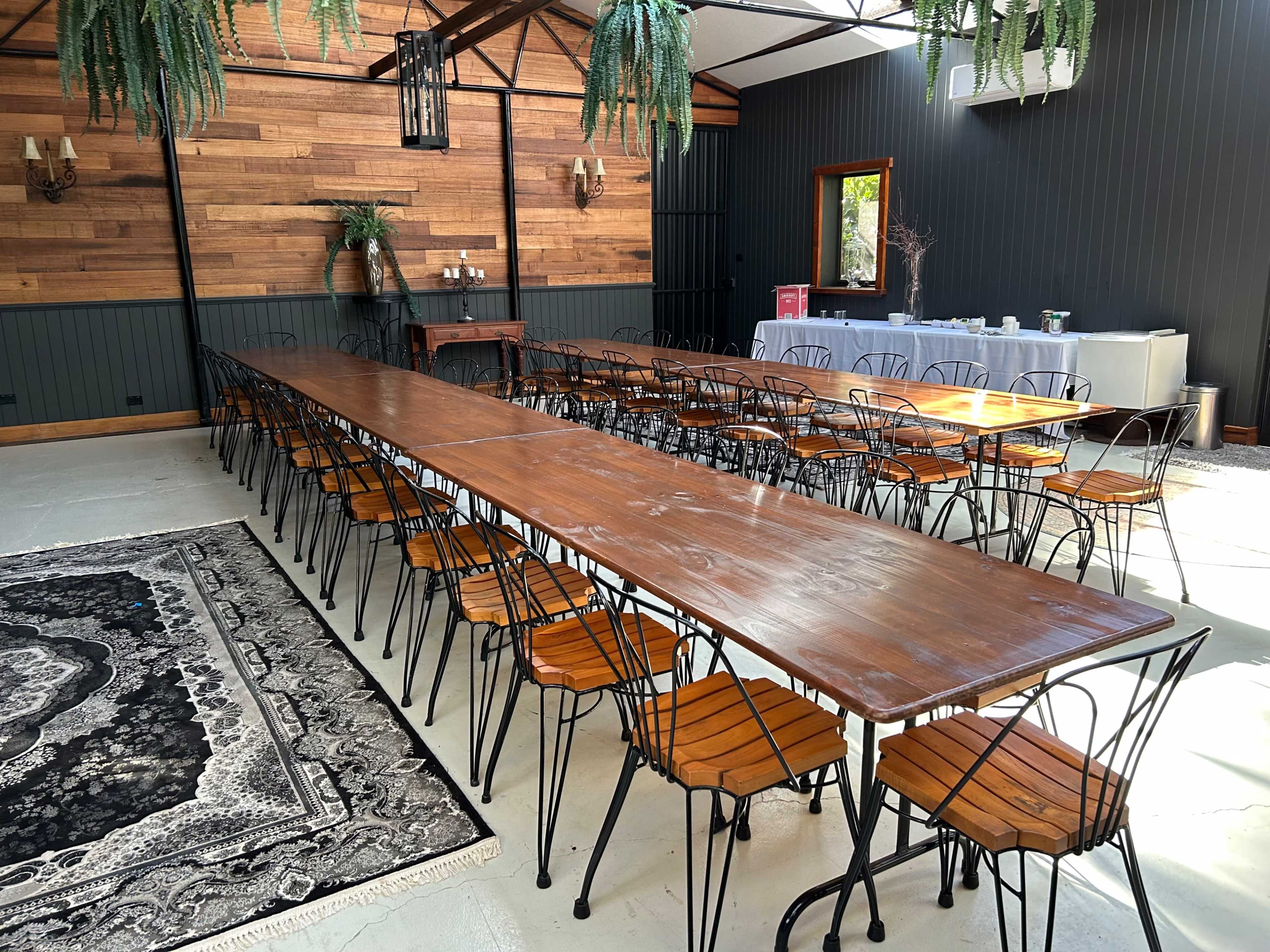 A long wooden dining table with metal chairs is set in a room with wooden walls and greenery, ready for an event.