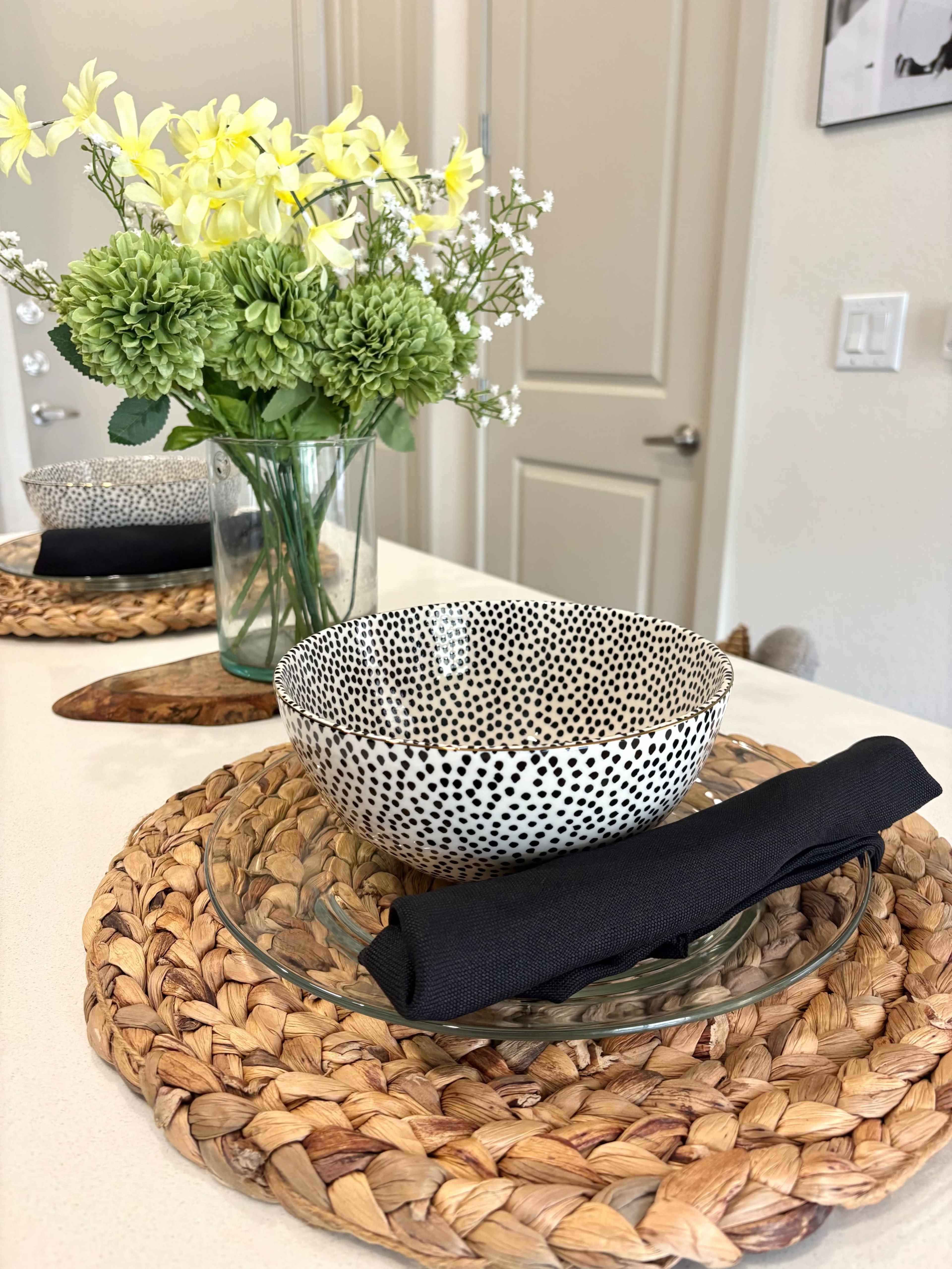 A patterned bowl sits on a glass plate, accompanied by a black napkin, beside a vase of green and yellow flowers on a woven placemat.
