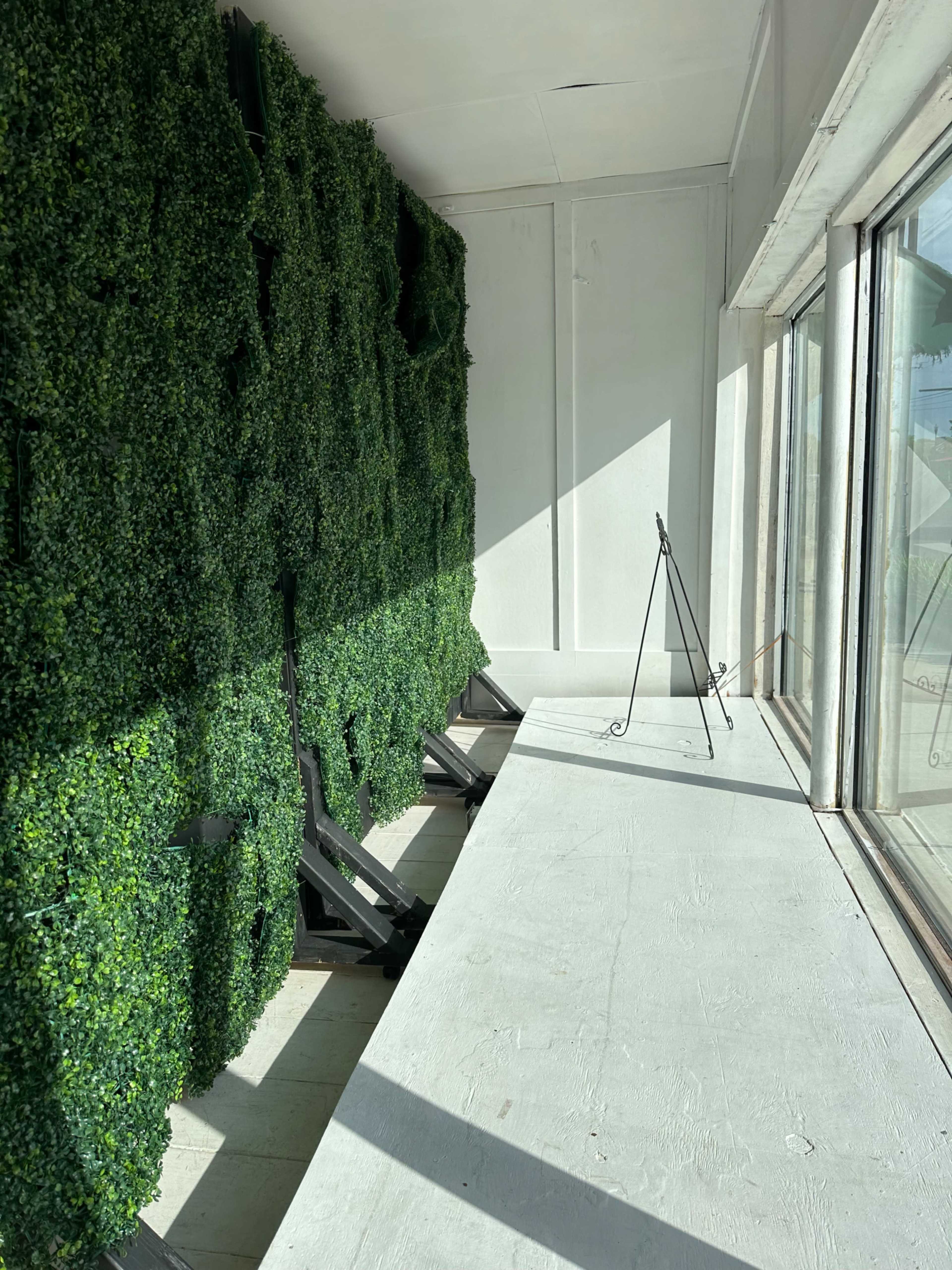 A sunlit indoor space featuring a green wall of artificial foliage alongside a long, empty table with a metal easel.