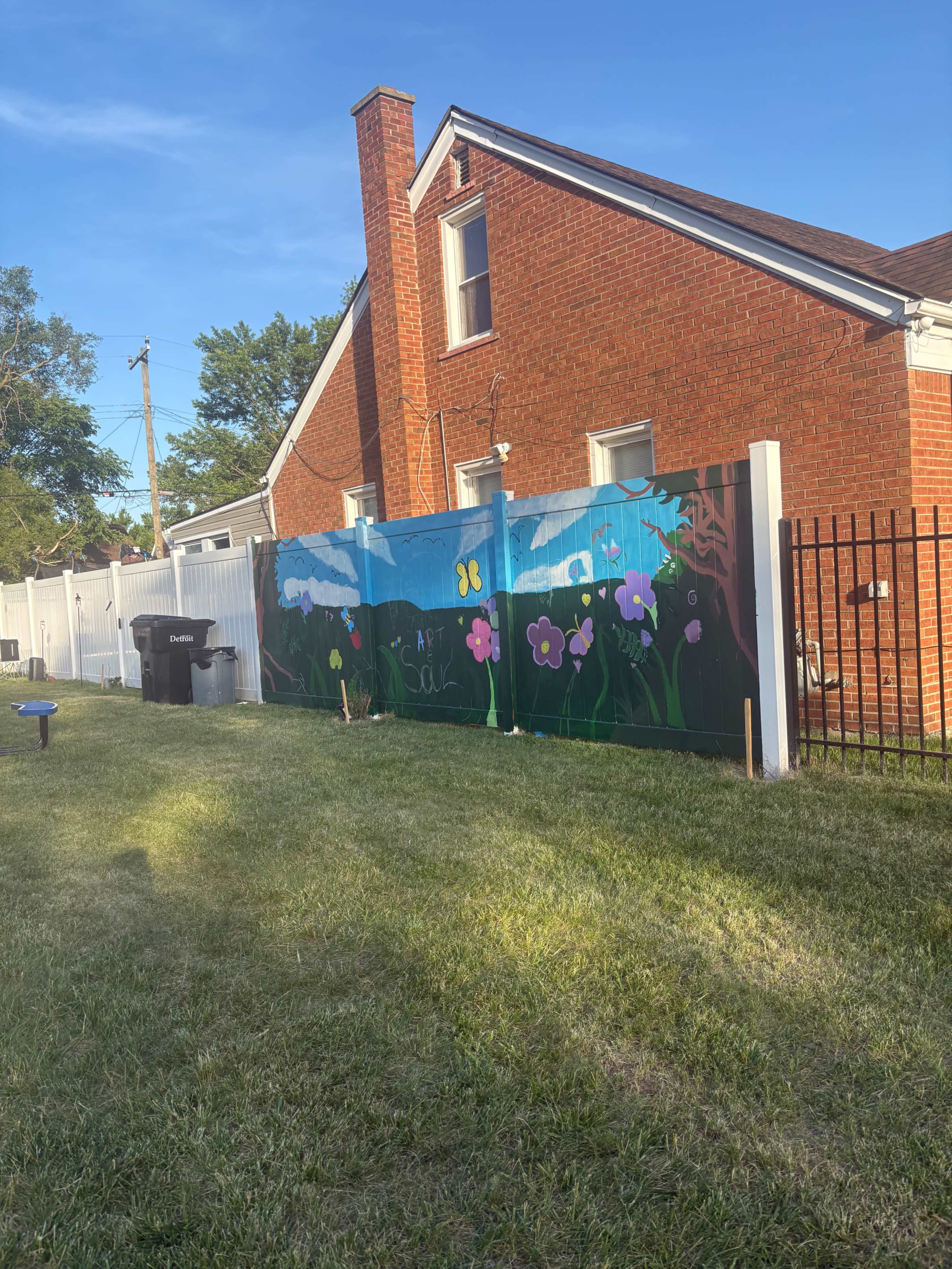 A colorful mural featuring flowers and butterflies is painted on a fence in a grassy backyard, next to a red brick house.