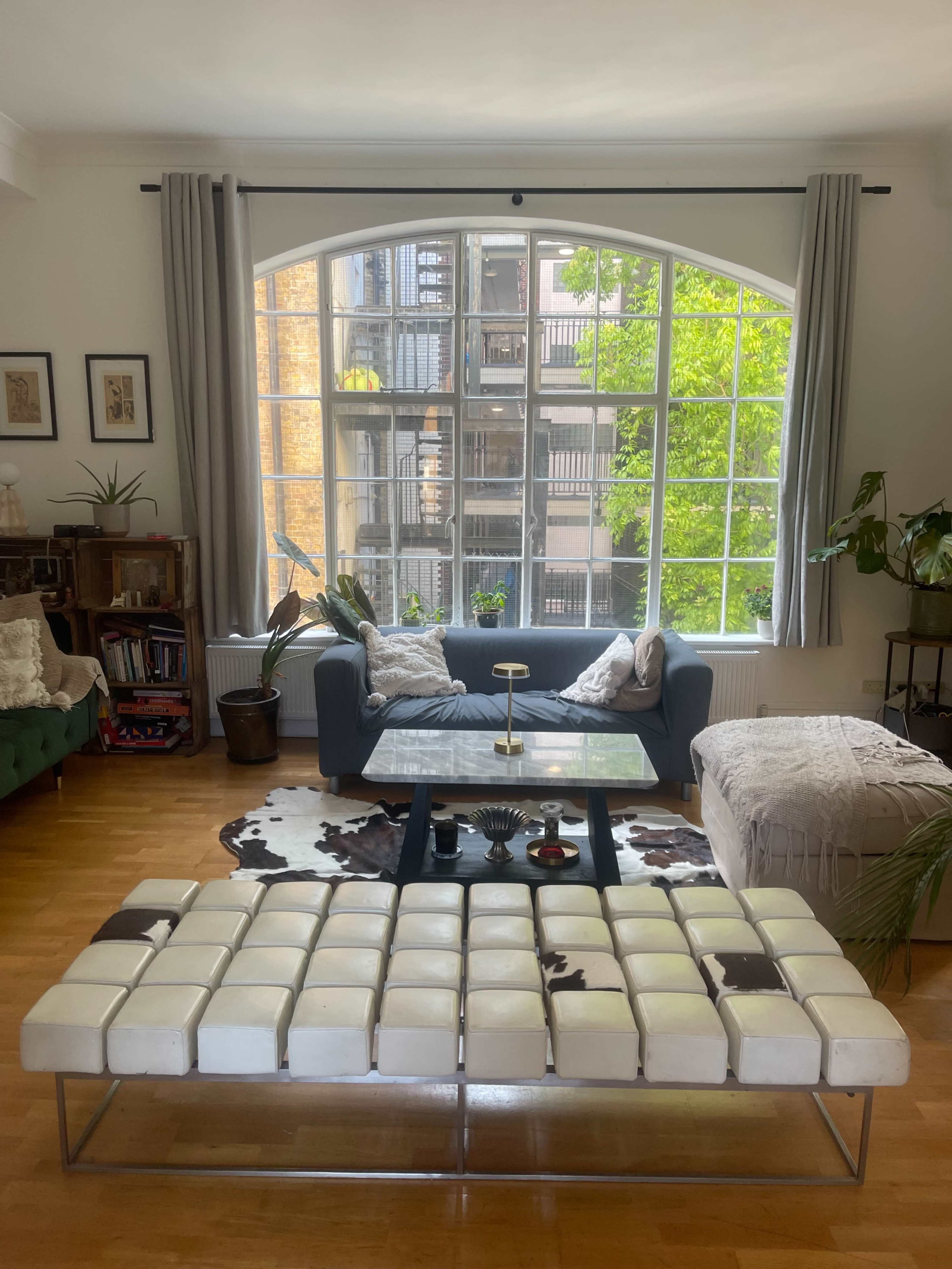 The image shows a cozy living room with a large arched window, a coffee table in the center, and various plants and furniture arranged around the space.
