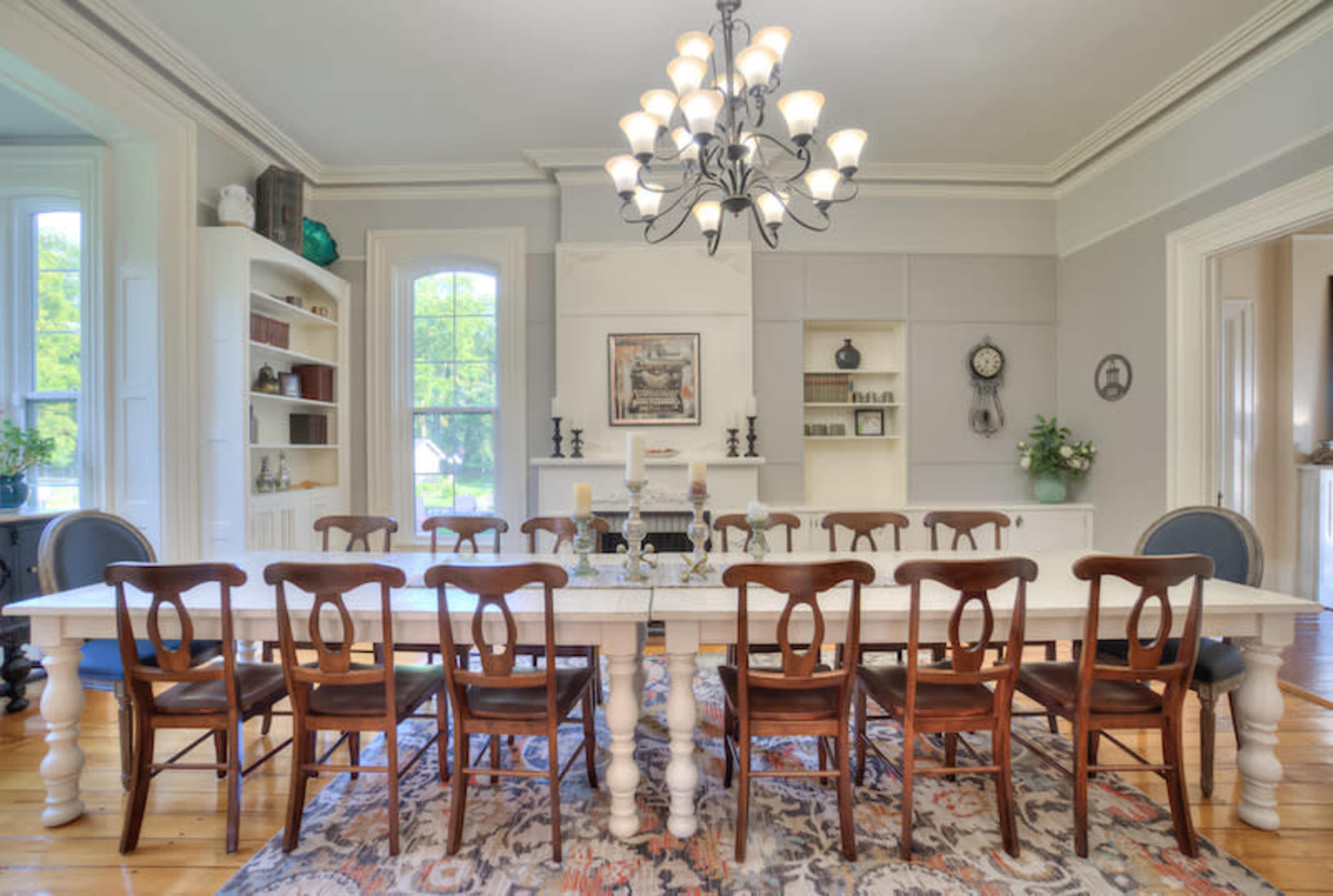 A long, white dining table surrounded by wooden chairs is set in a well-lit room with large windows and decorative shelving.