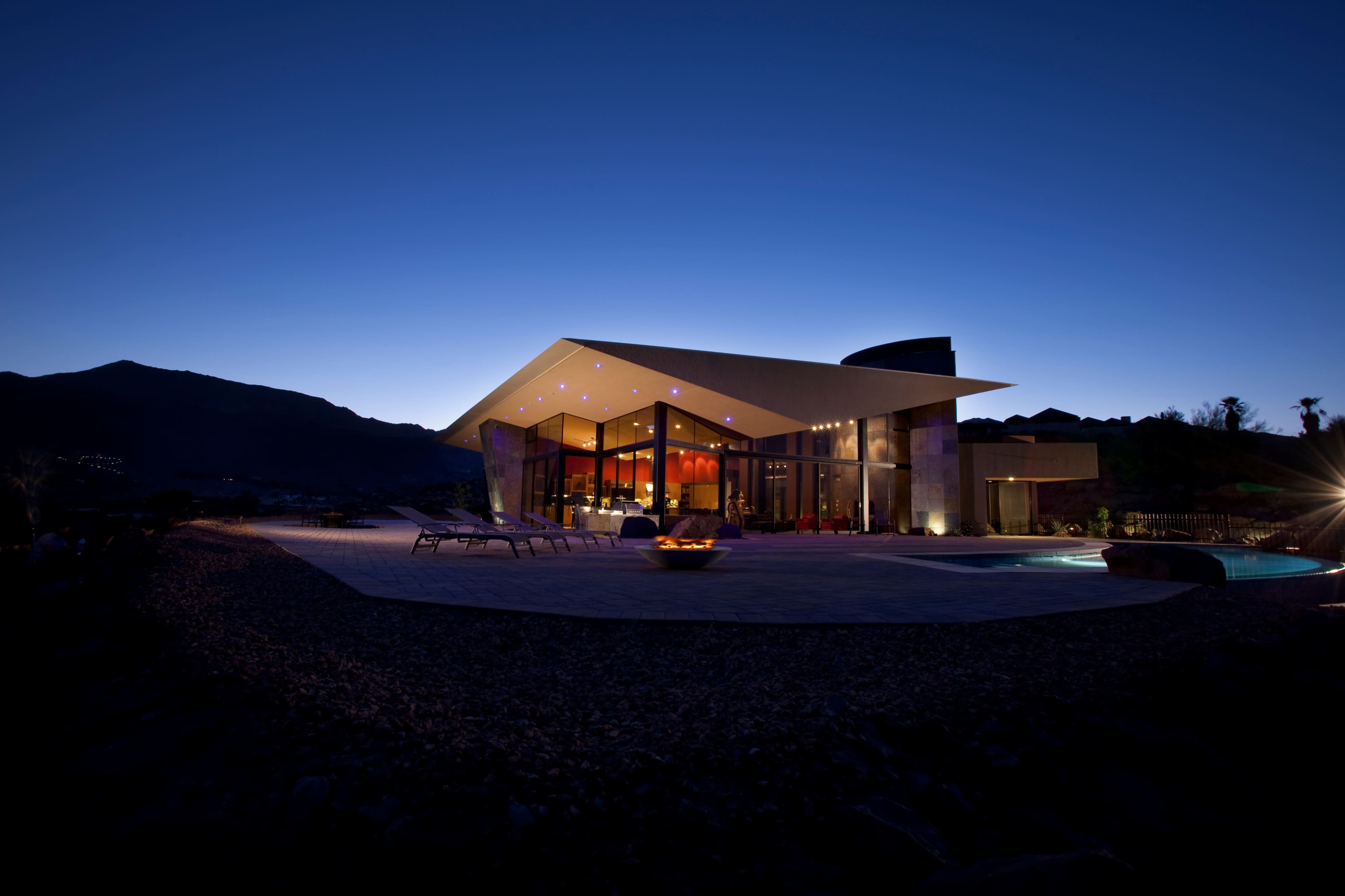 A modern architectural villa with large glass windows and a flat roof, illuminated at twilight against a mountainous backdrop.