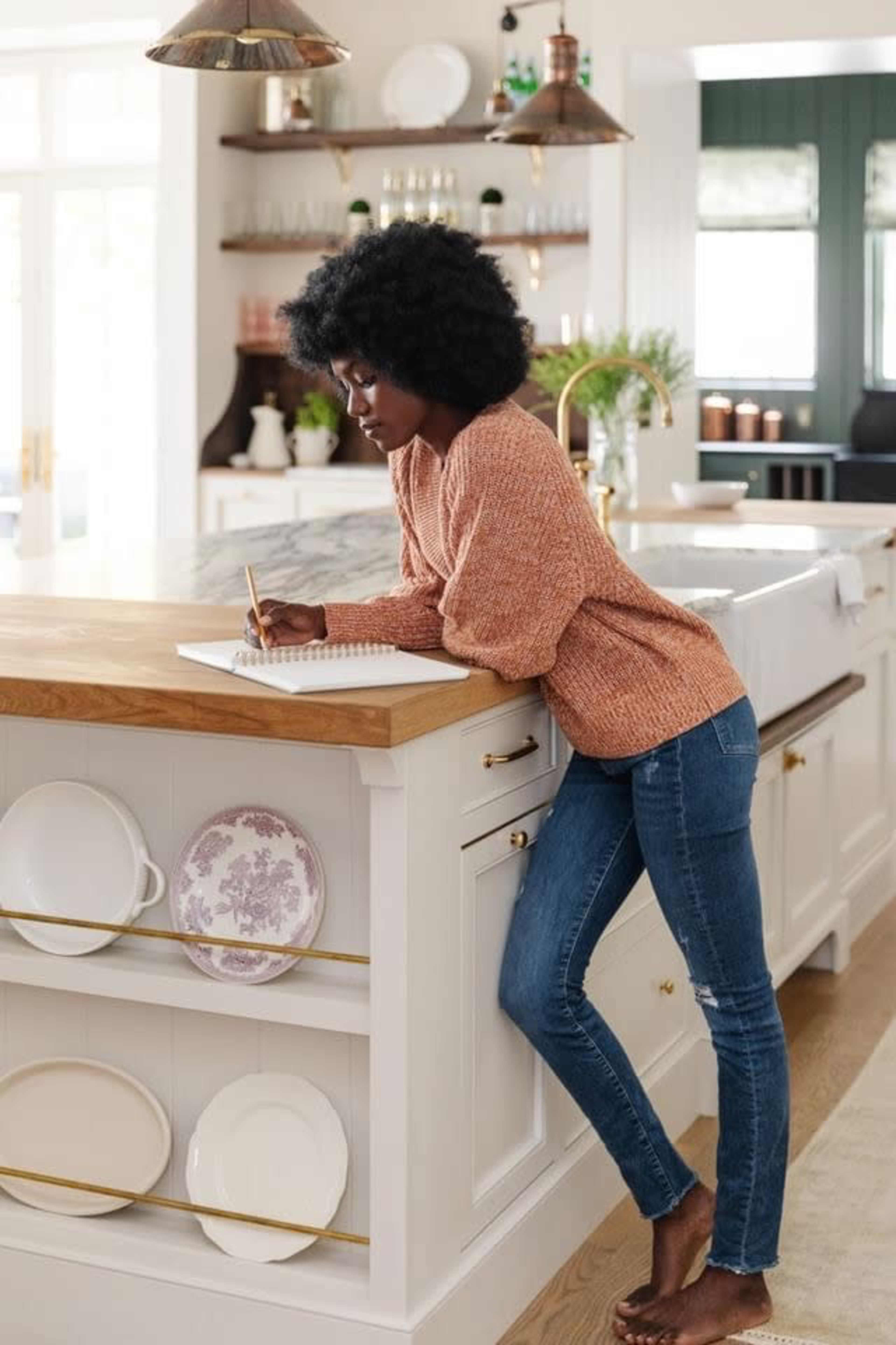 A woman writes in a notebook while leaning on a kitchen island in a modern kitchen.