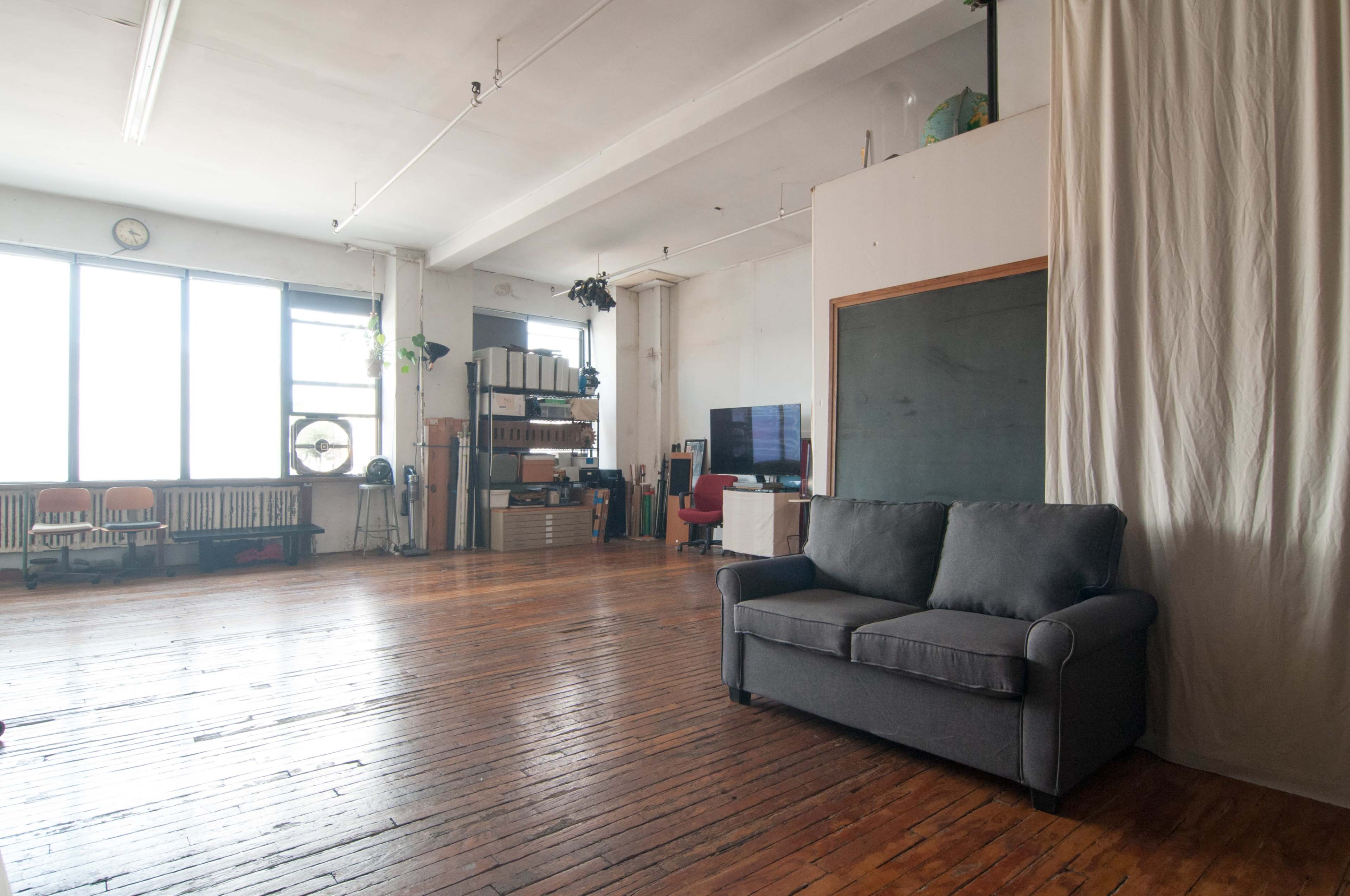 The image shows a spacious room with wooden floors, large windows allowing natural light, and a gray sofa positioned near a wall draped with fabric.