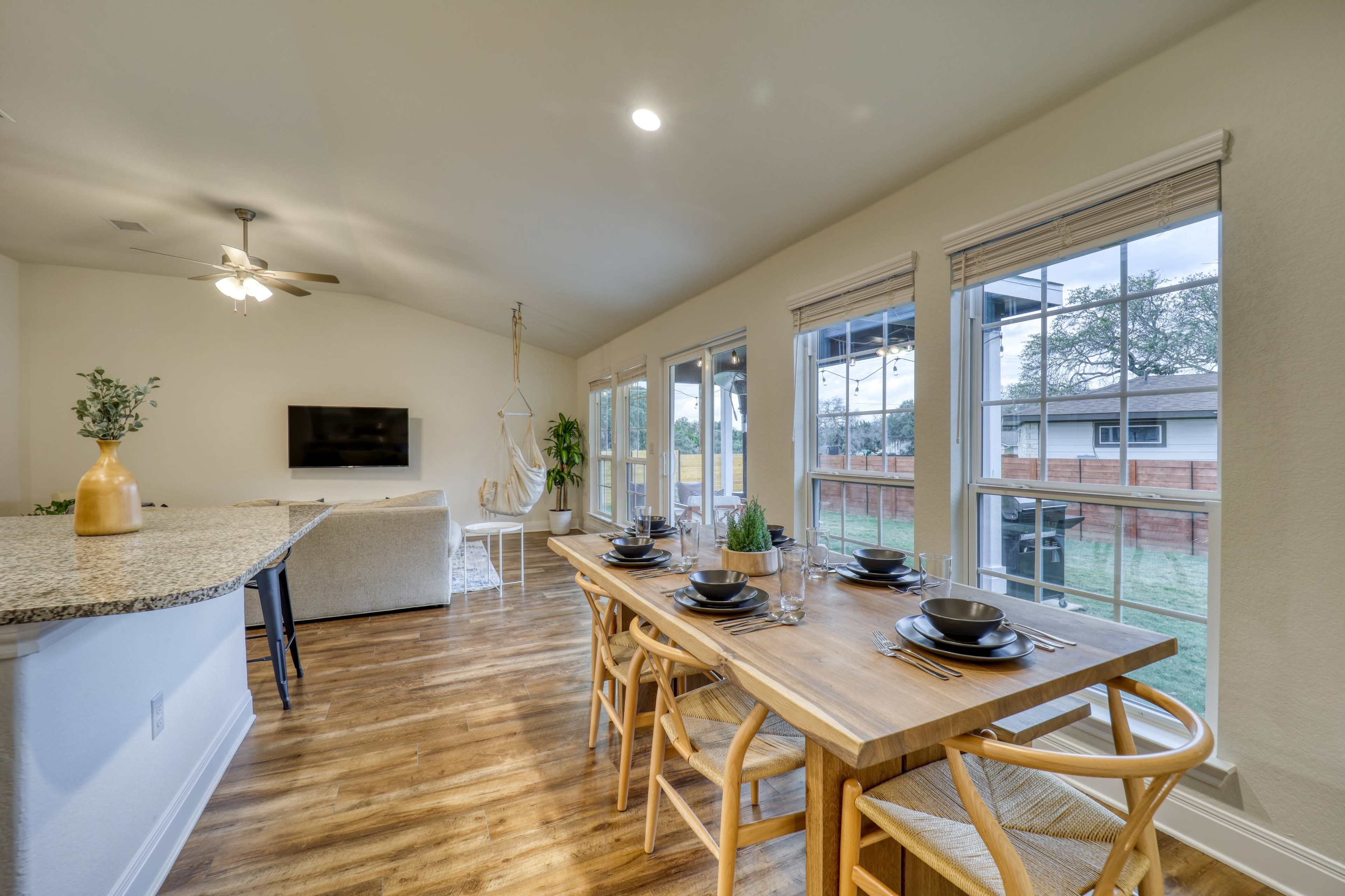 A dining area featuring a wooden table set with plates and cutlery, adjacent to a living space with a television and large windows overlooking a yard.