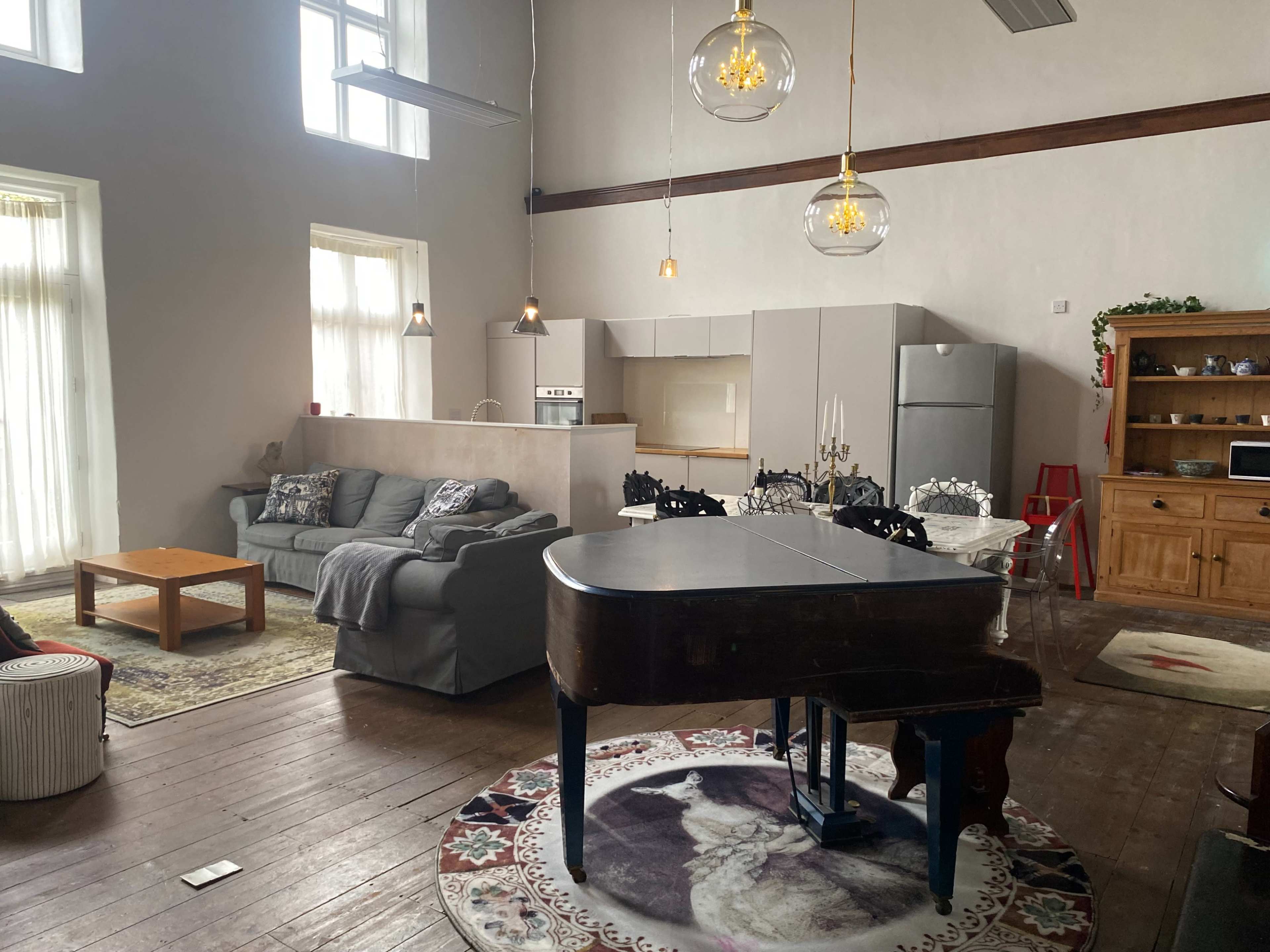 A spacious living area featuring a grand piano, a gray sofa, and a kitchen in the background with modern appliances.