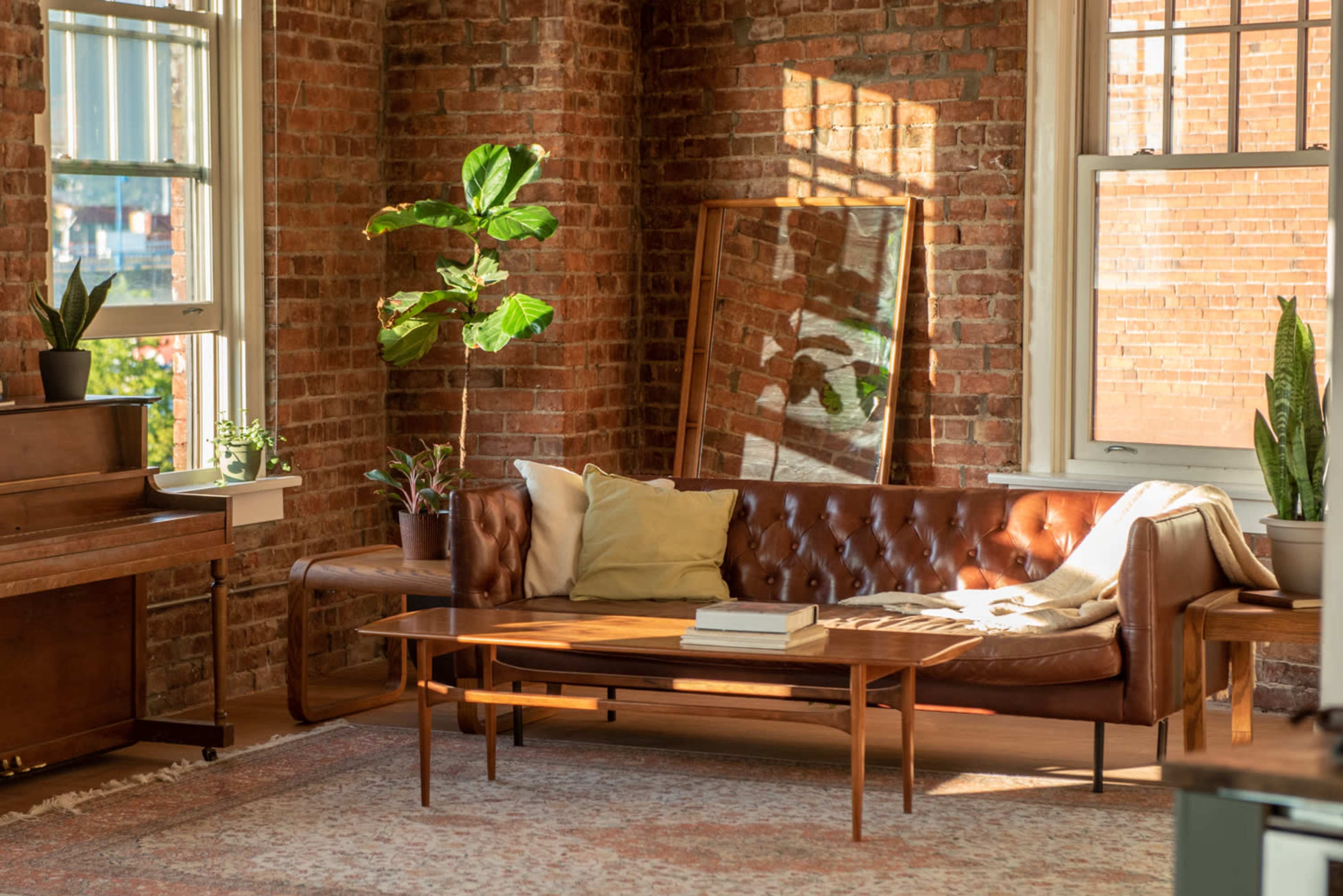 A living room with a brown leather couch, a wooden coffee table, and large windows allowing natural light to illuminate the space, which features exposed brick walls and various indoor plants.