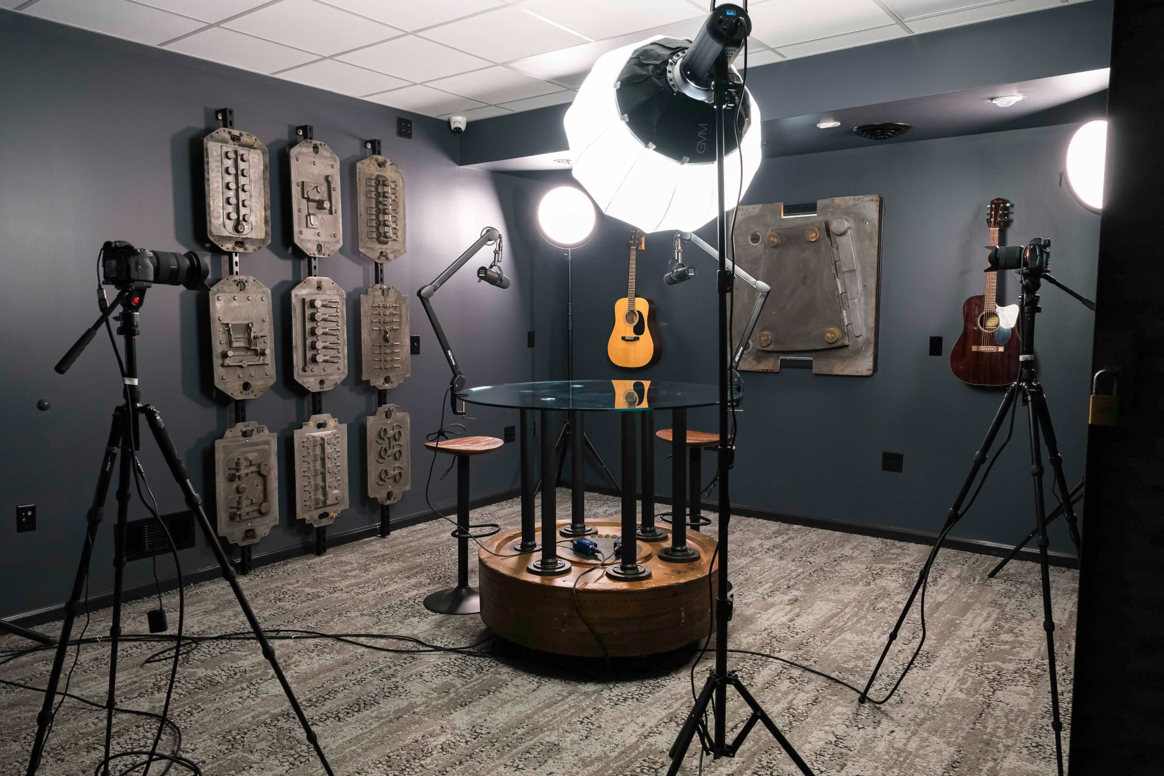 A photography studio with a circular glass table, two guitars mounted on the wall, and multiple cameras set up on tripods under soft lighting.