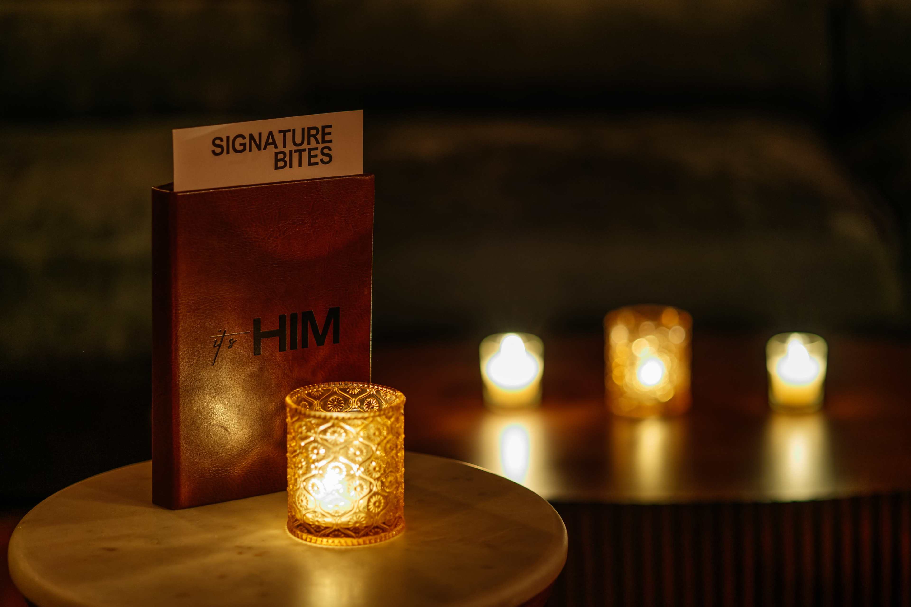 A menu titled "Signature Bites" is placed on a small table beside lit candles in decorative holders.