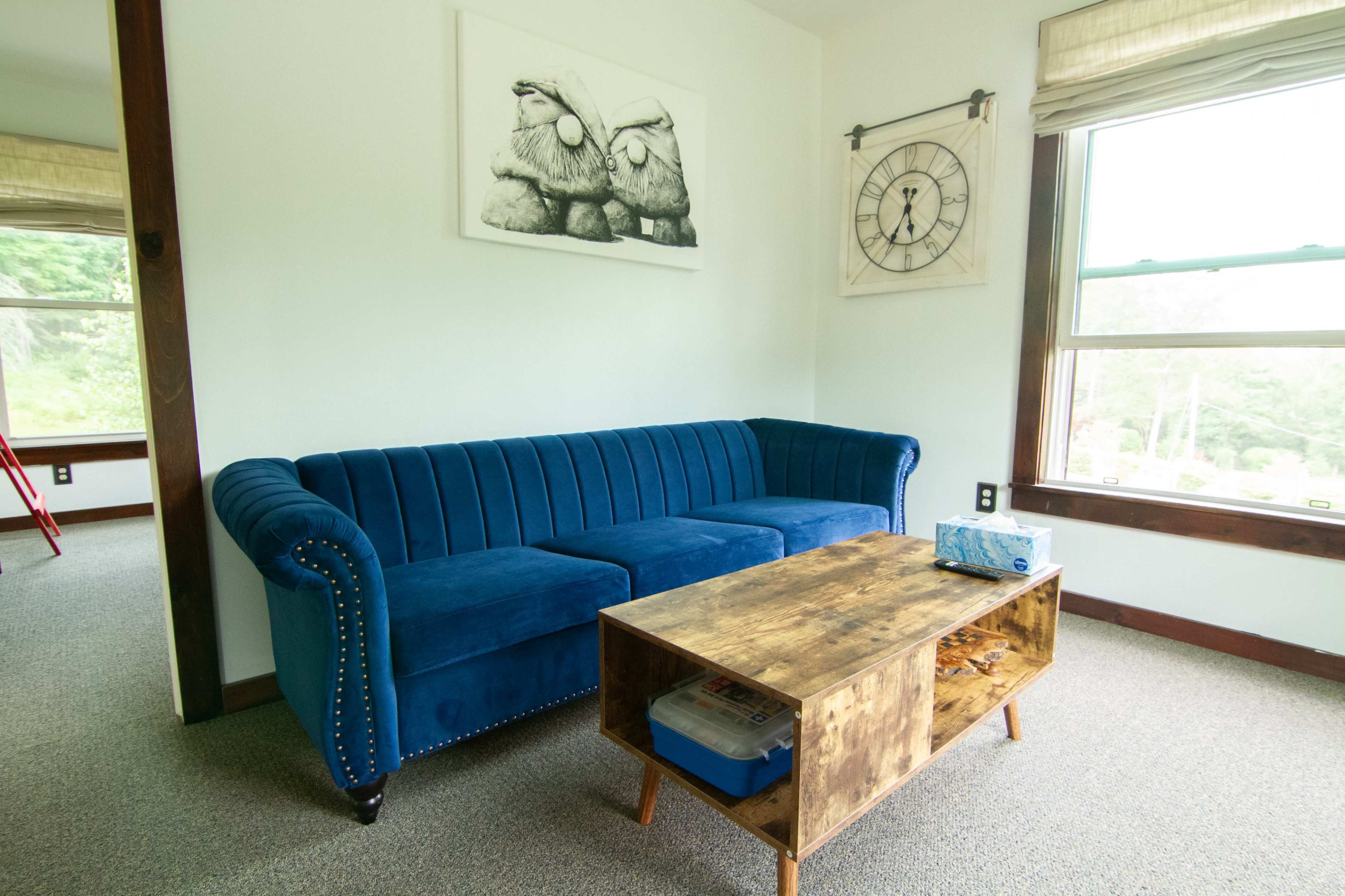 The image shows a blue velvet couch and a wooden coffee table in a bright, minimalist living room with large windows.