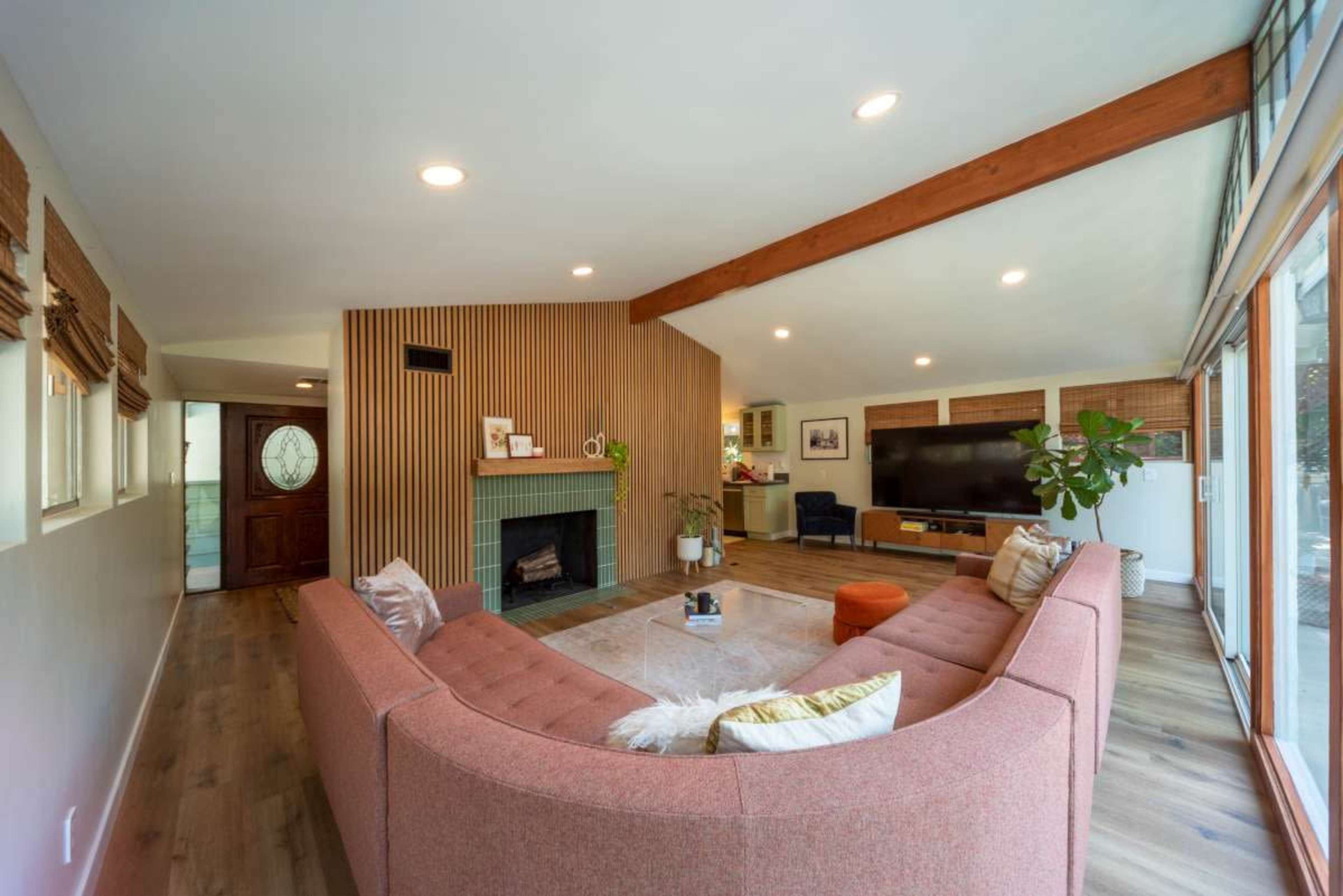 A modern living room featuring a pink sectional sofa, a fireplace with wooden paneling, and large windows with natural light.