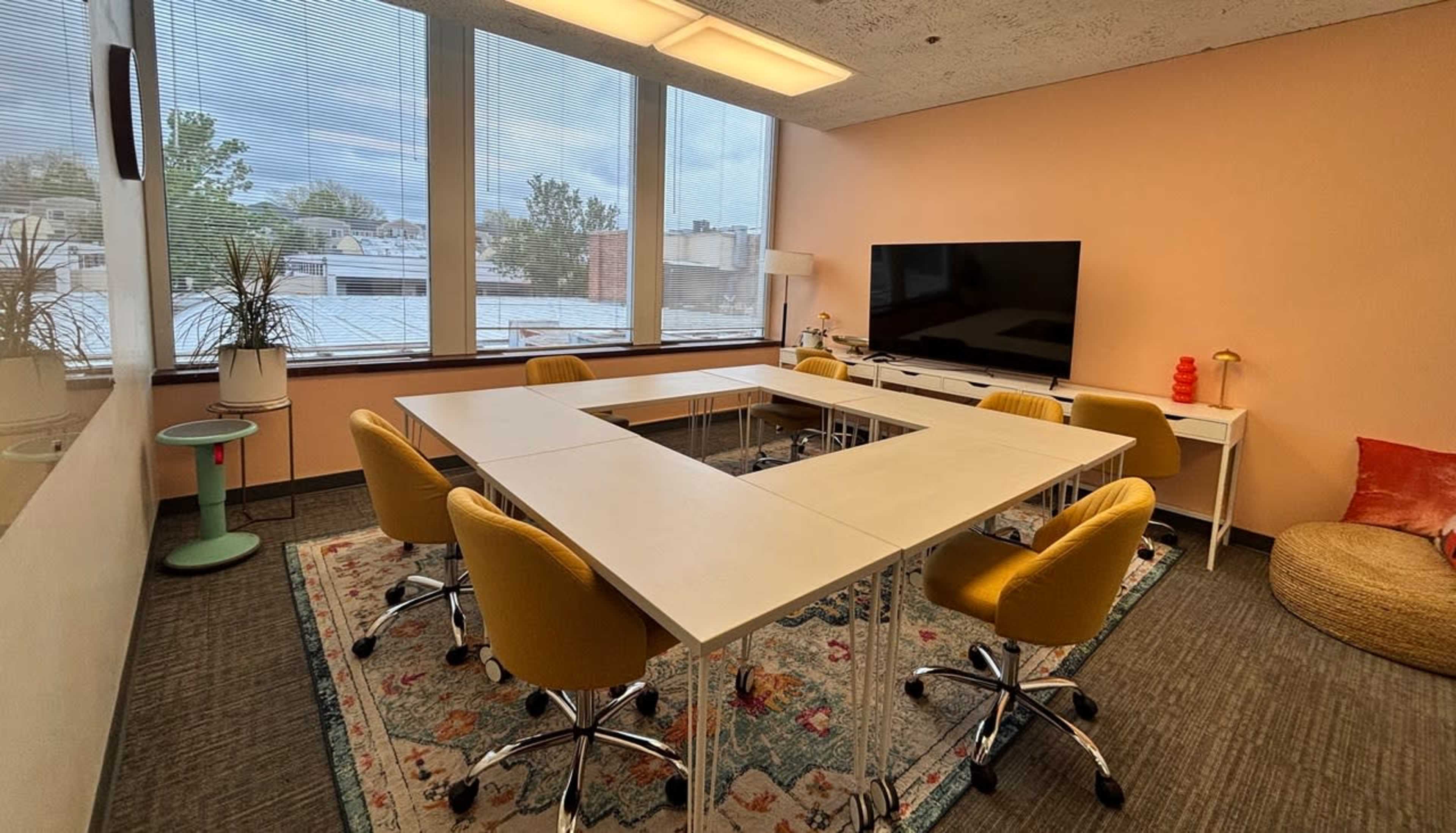 A meeting room features a rectangular arrangement of white tables surrounded by yellow chairs, with a large TV and a colorful rug on the floor.