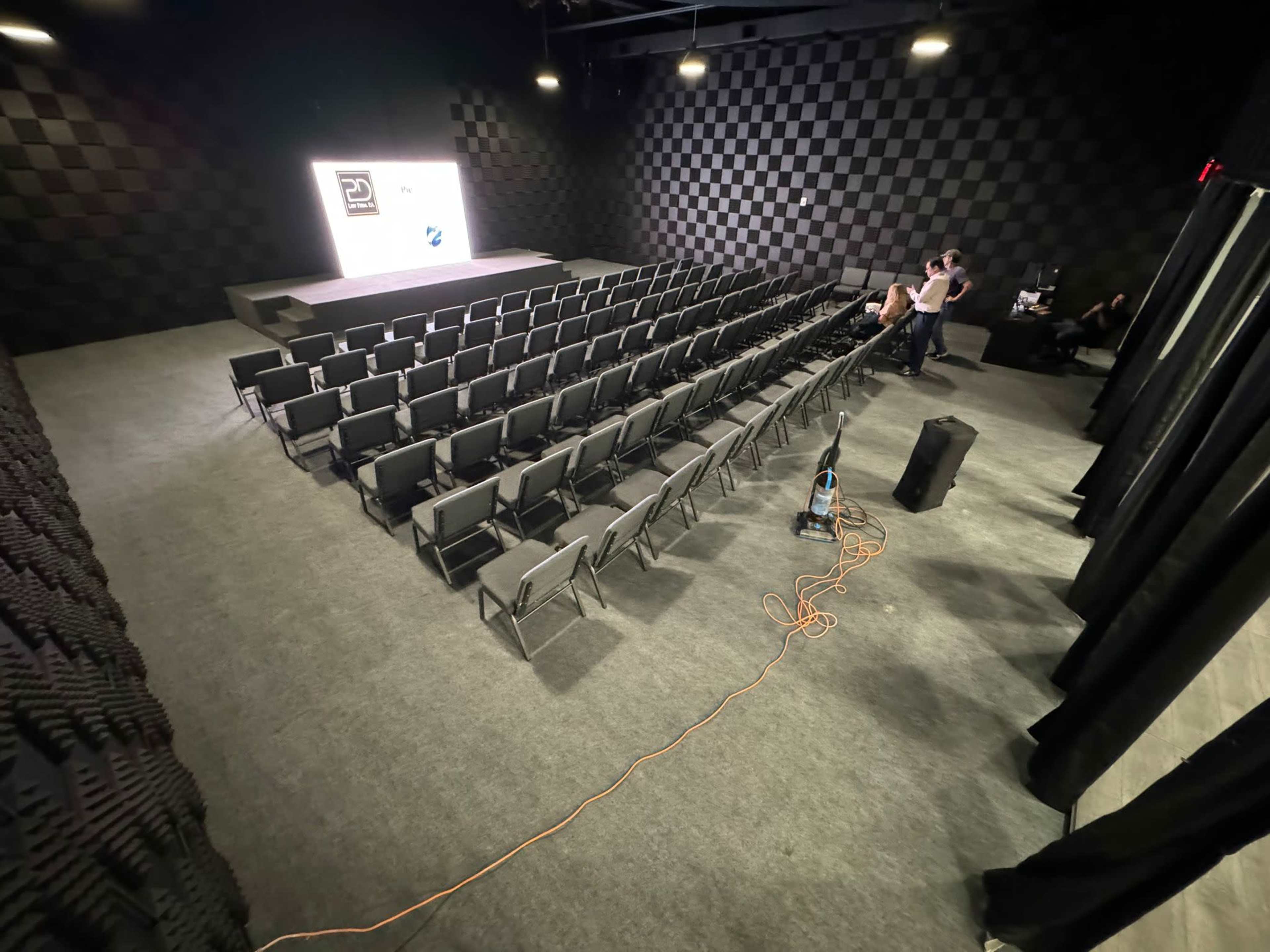The image shows a black and white checkered auditorium with rows of chairs arranged in front of a presentation screen.