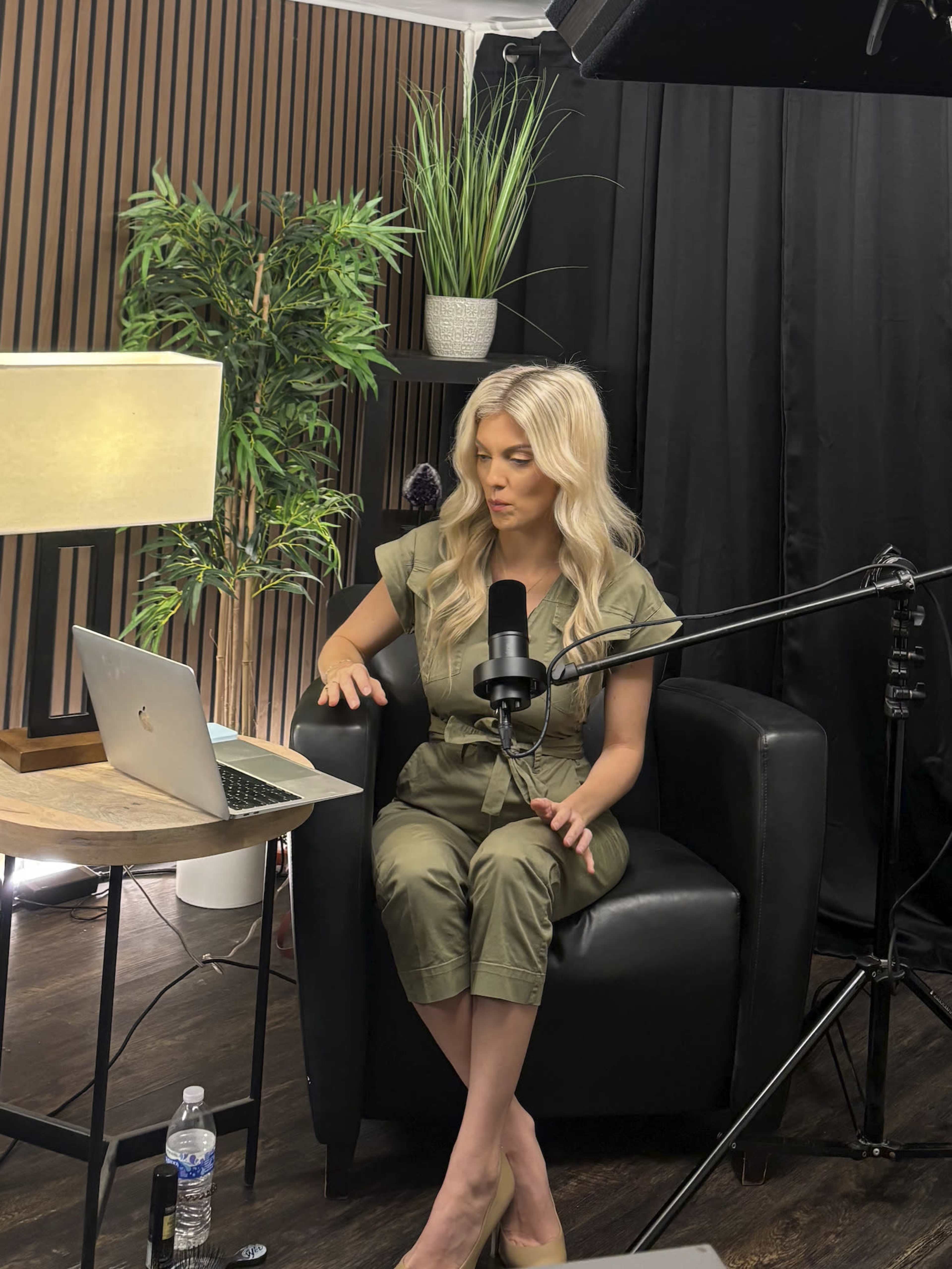 A woman in a green jumpsuit sits in a chair in a studio setting, speaking into a microphone while looking at a laptop on a nearby table.