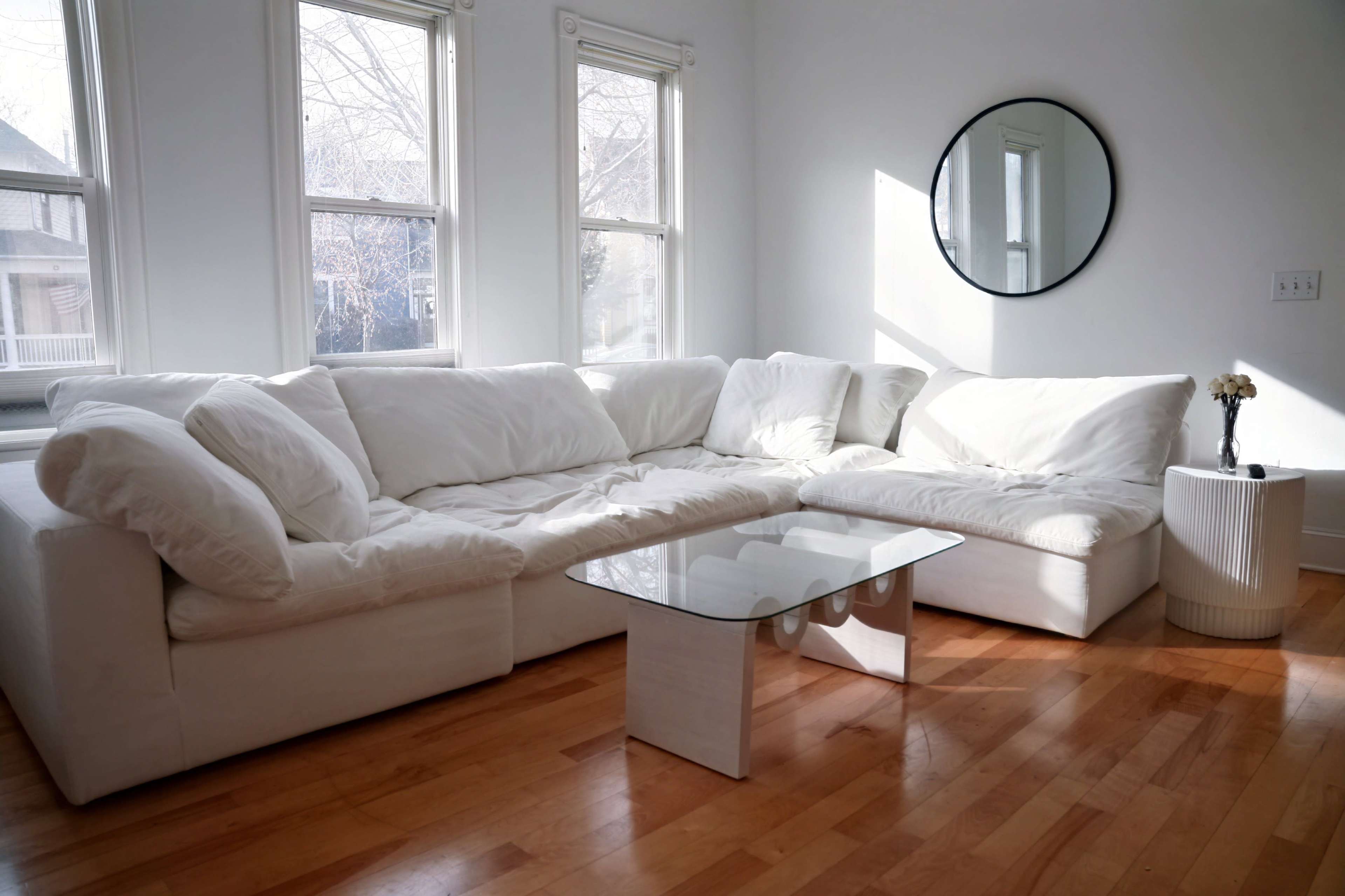 A bright room with a white sectional sofa, a glass coffee table, and large windows allowing natural light to enter.