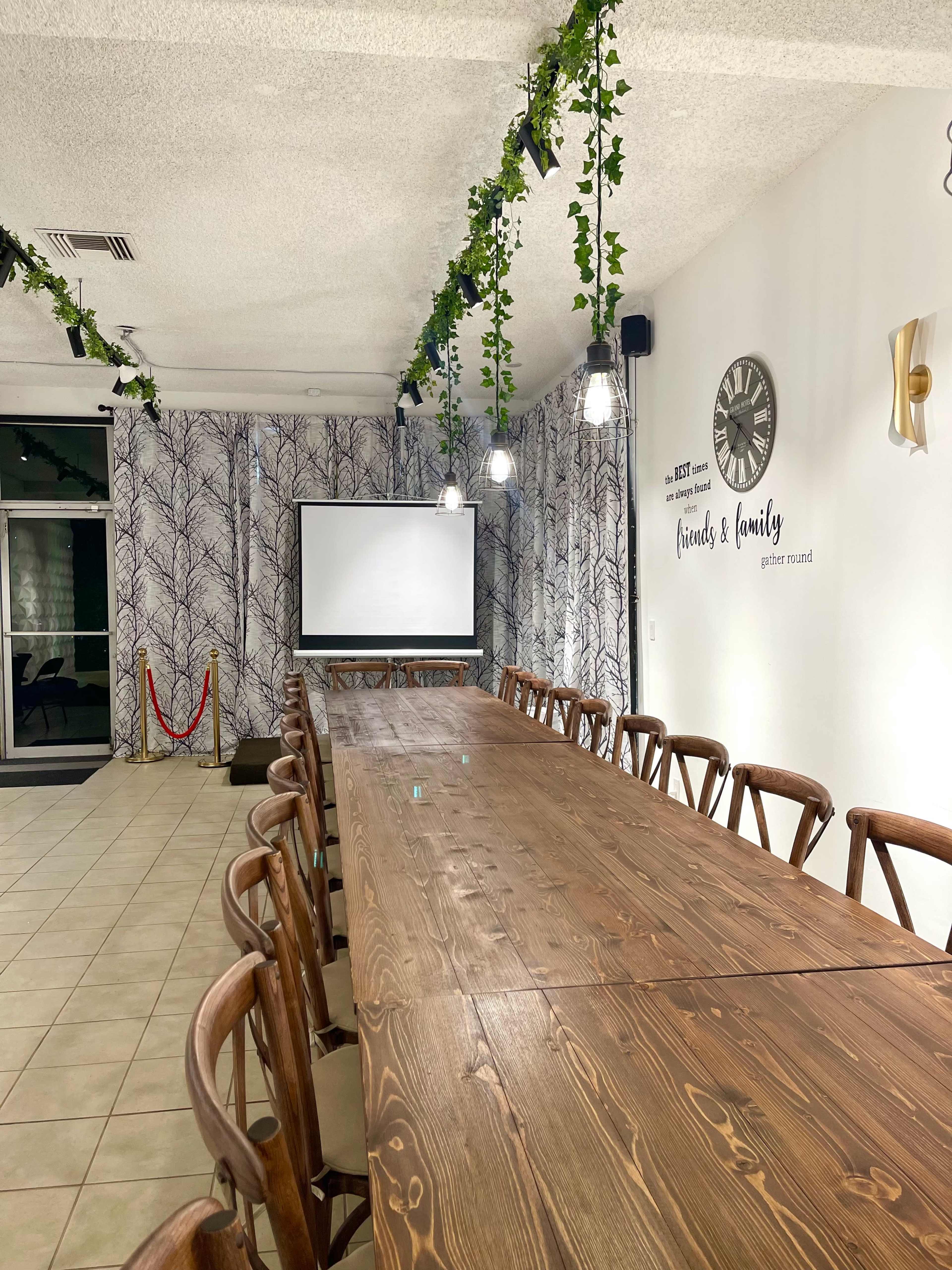 A long wooden table surrounded by chairs in a well-lit meeting room, featuring a projector screen and decorative wall art.