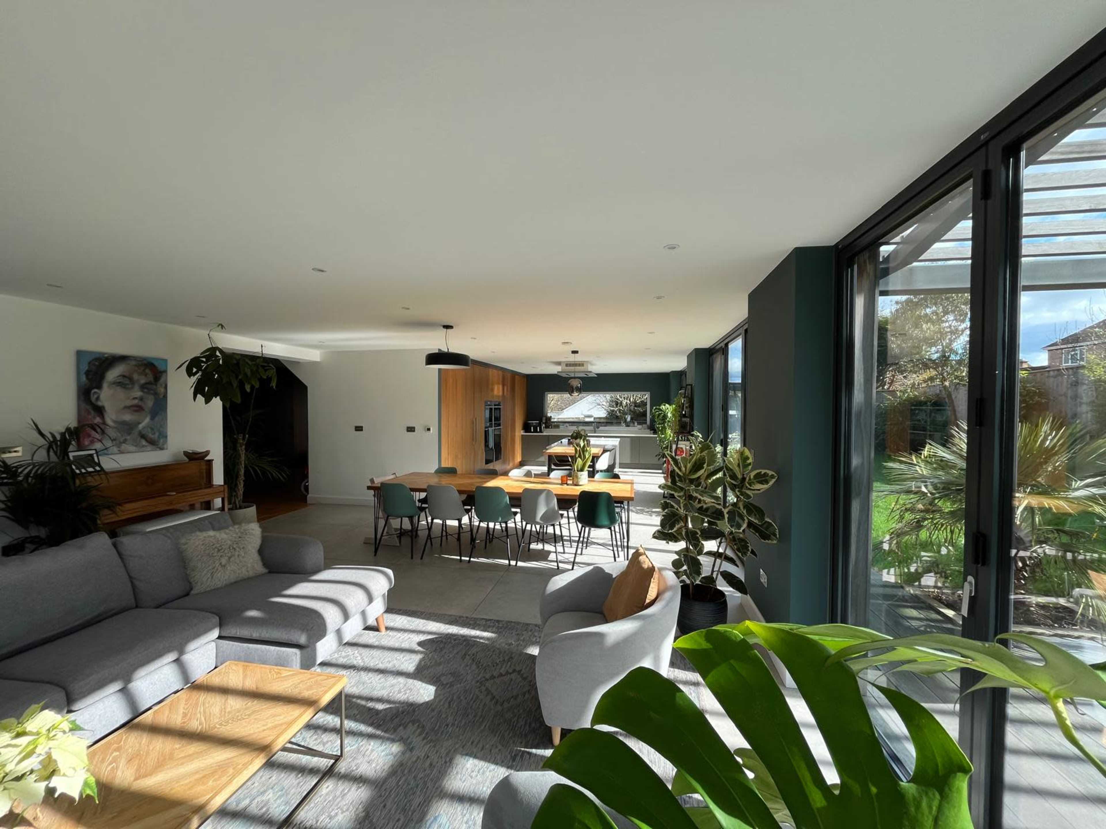 A modern living space features a large sofa, a dining area with multiple chairs, and large glass doors opening to an outdoor garden.