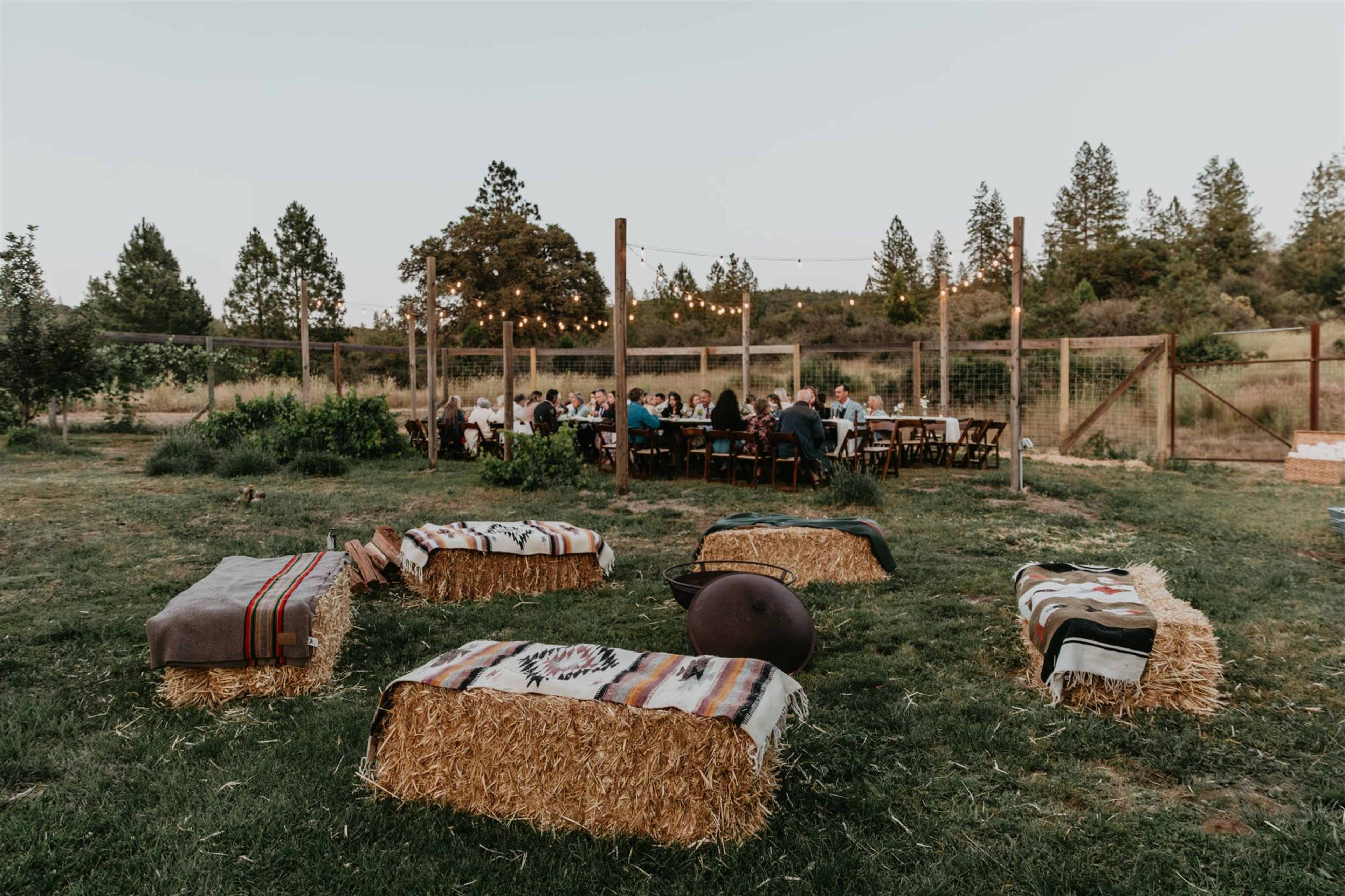 A rustic outdoor gathering features a long table set for dining, surrounded by straw bales used as seating, beneath string lights and trees.