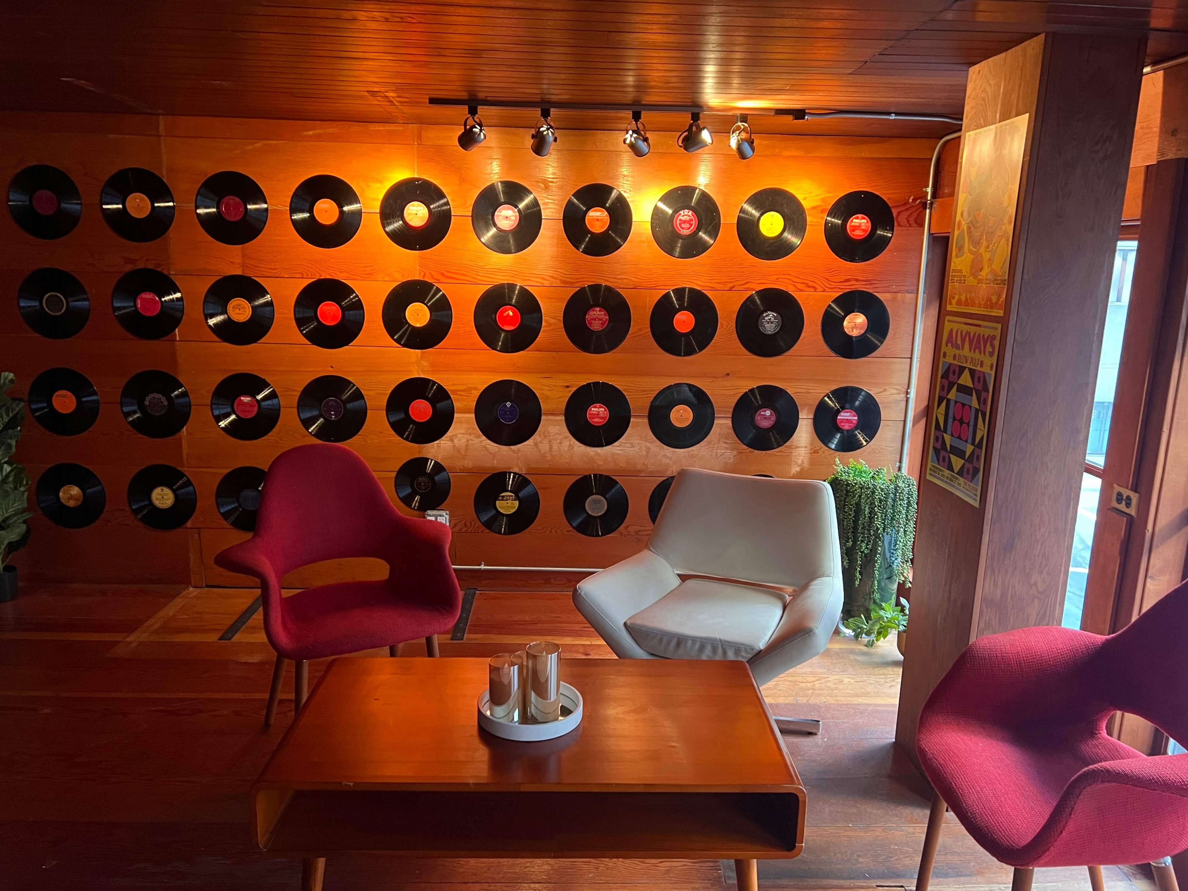 A room with a wooden wall adorned with various vinyl records, accompanied by two modern chairs and a coffee table in a cozy seating area.