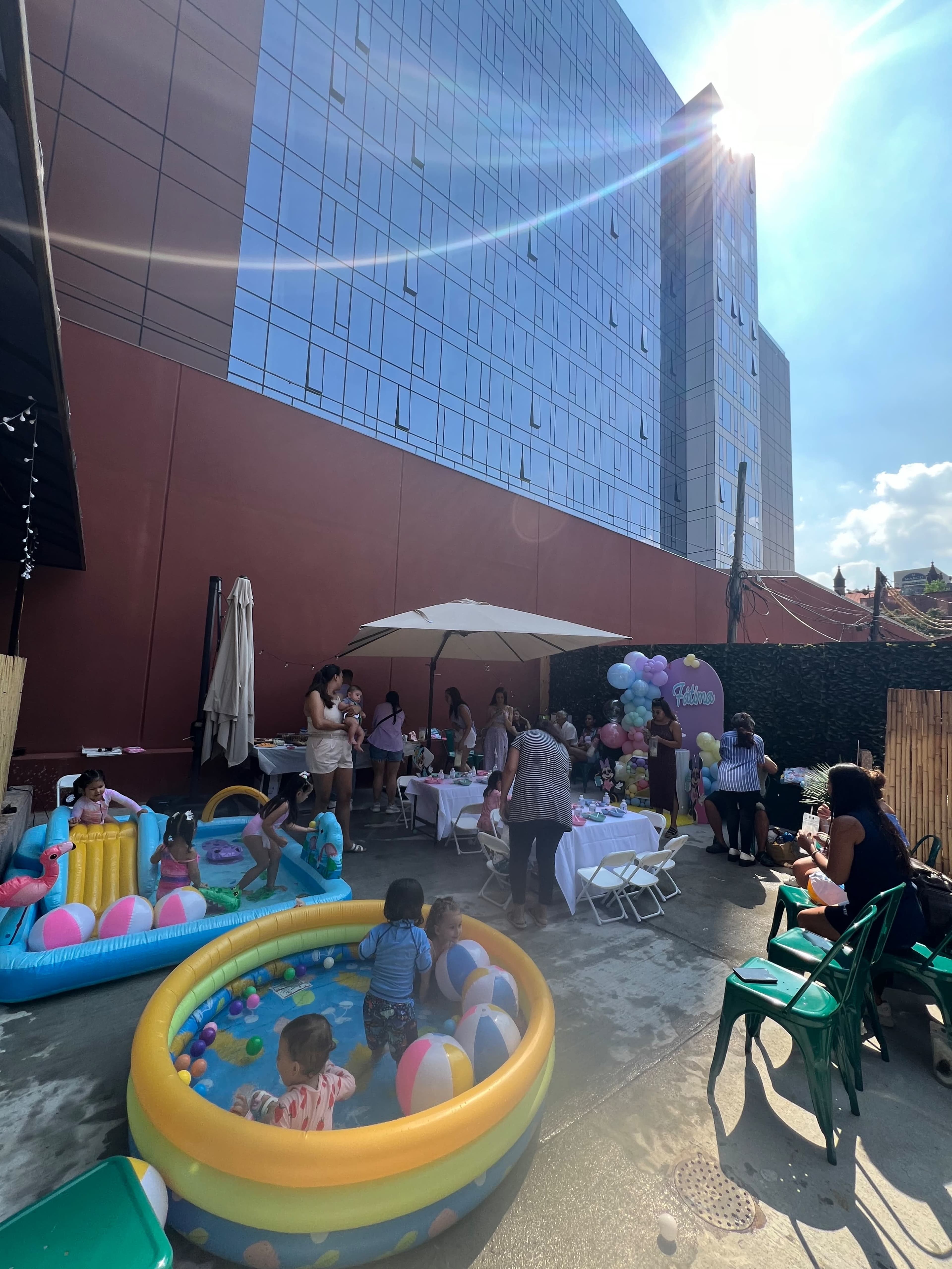 The scene shows a backyard party with children playing in inflatable pools and adults gathered around tables, while a large building looms in the background.