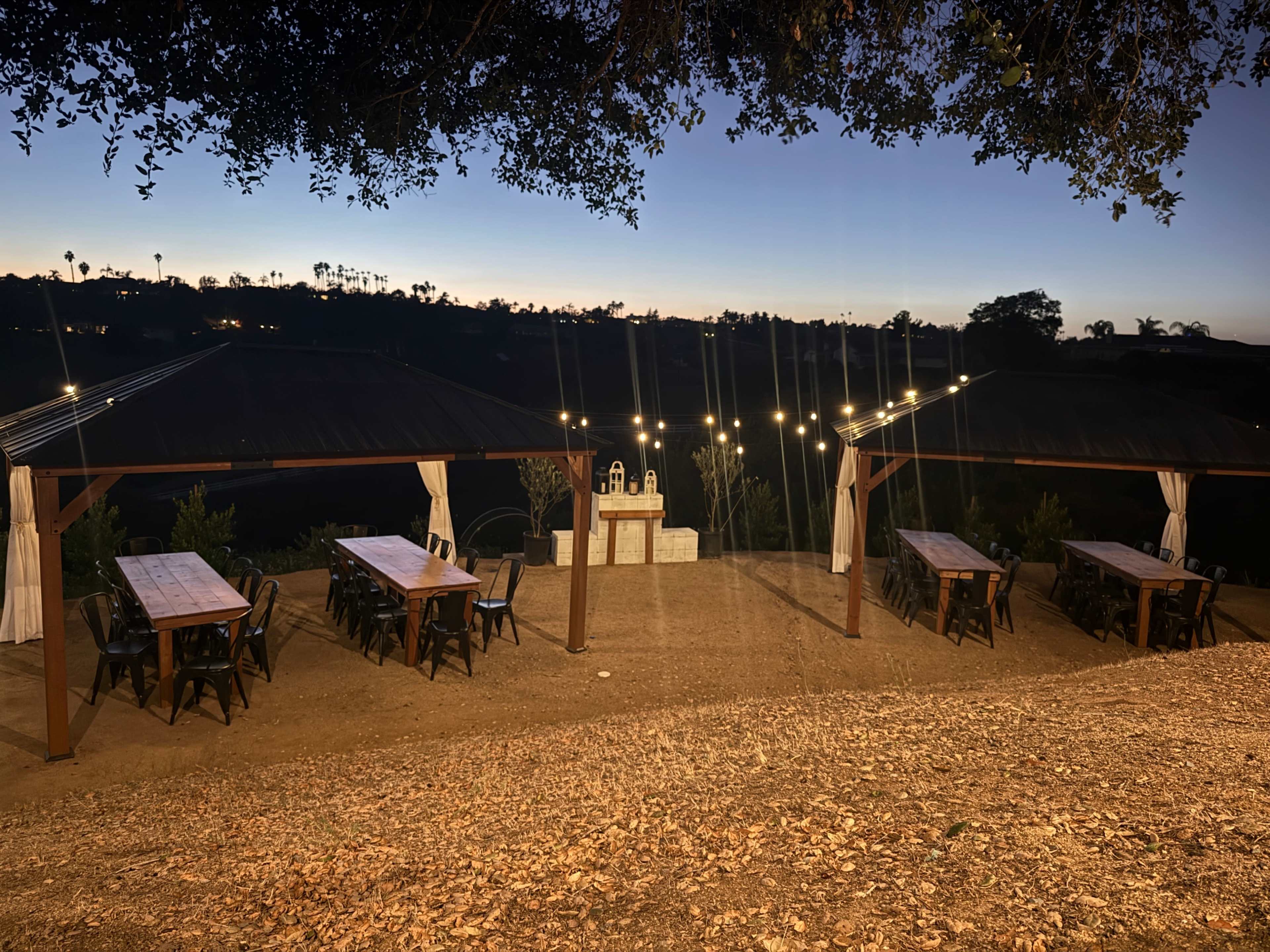 The scene shows a rustic outdoor dining area with several long tables and string lights, set against a twilight backdrop.