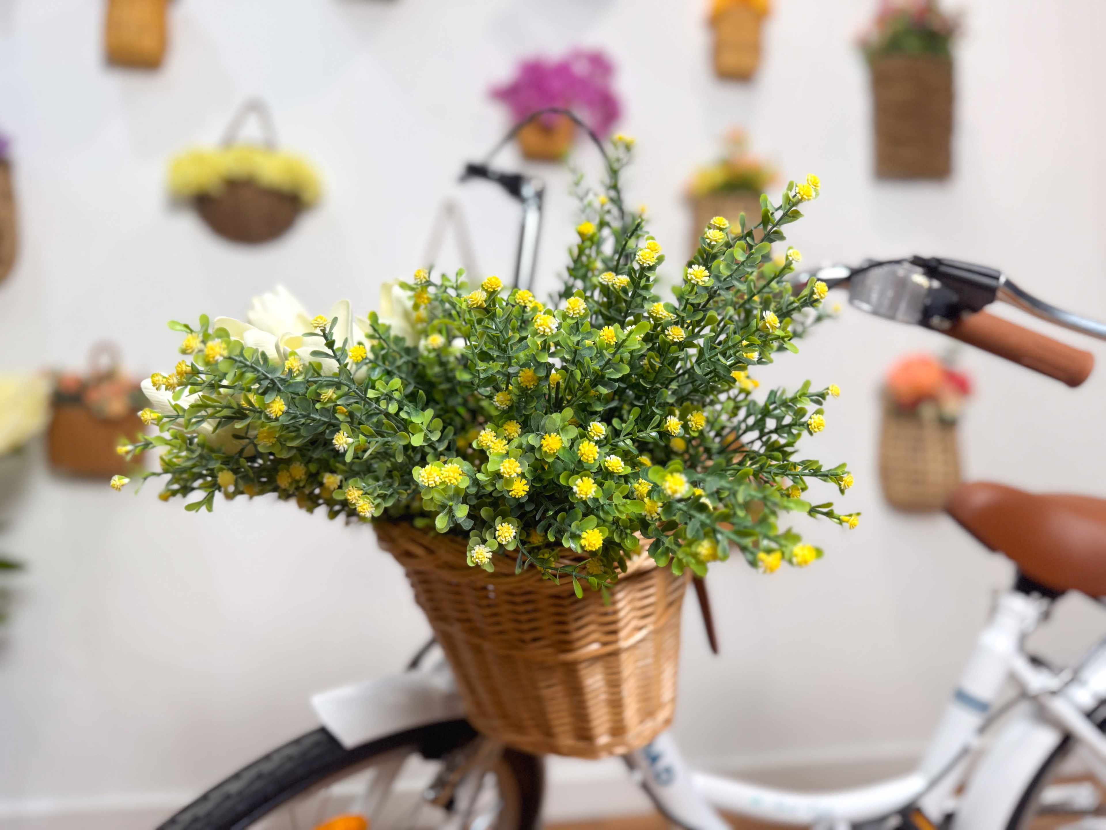 A bicycle with a woven basket is filled with bright yellow flowers, set against a backdrop of flower-filled wall baskets.