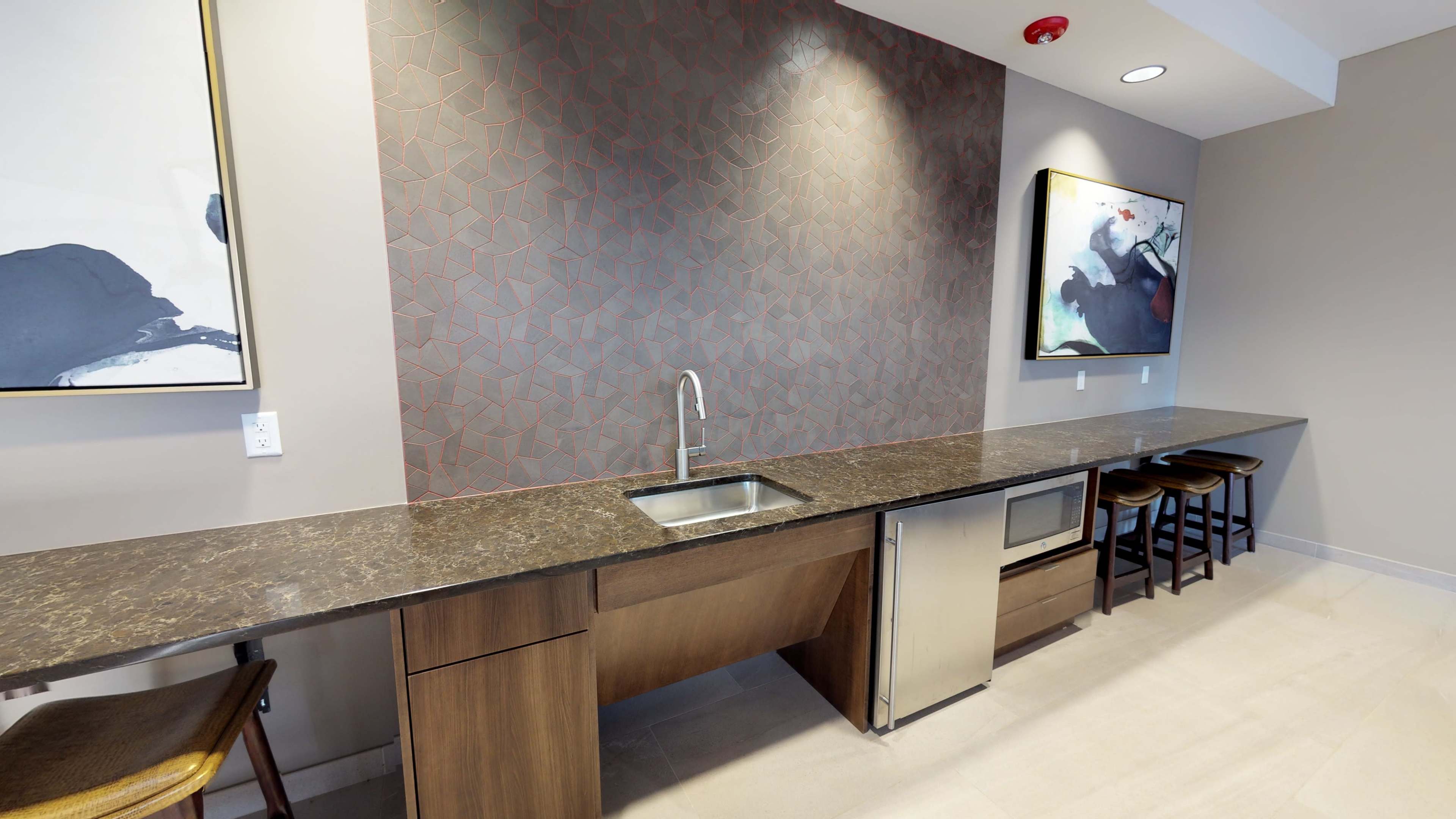 A modern kitchen features a dark countertop, a stainless steel sink, and a refrigerator, against a patterned wall with framed artwork above.
