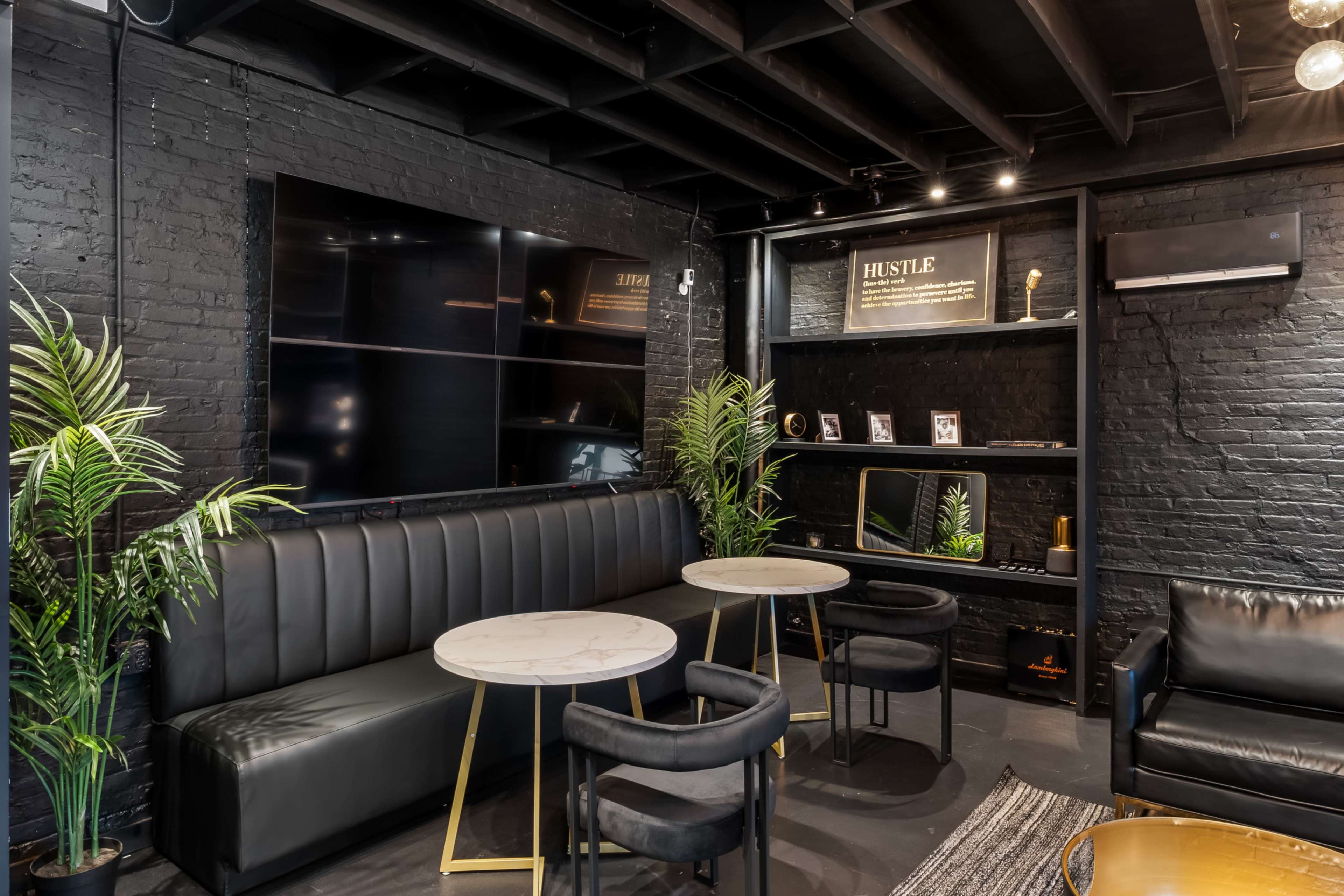 The image shows a modern lounge area with black walls, two round marble-top tables, and a shelf displaying framed photos and decorative items, accompanied by green plants and stylish seating.