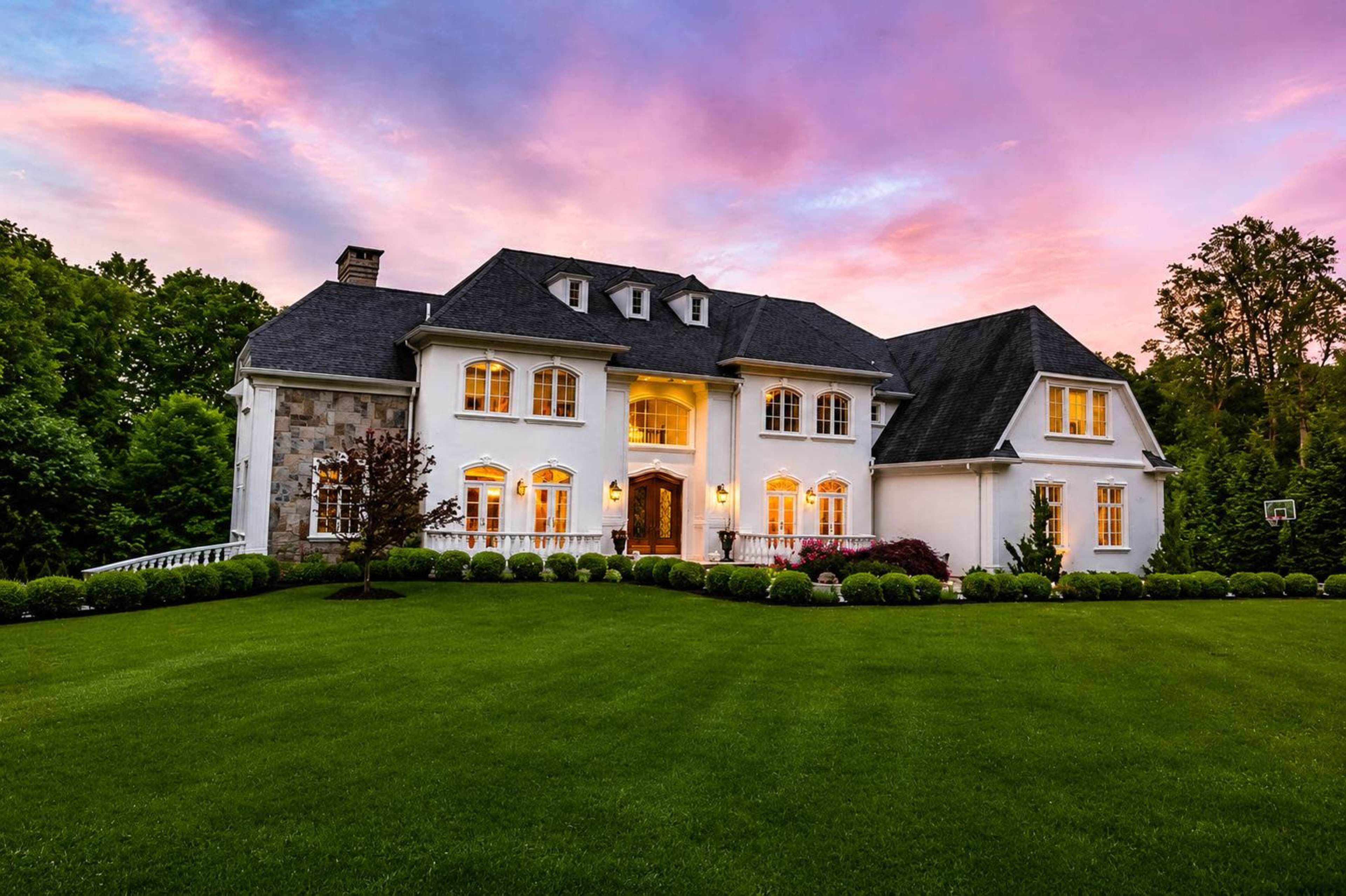 A large, two-story white house with multiple windows and a neatly manicured lawn is illuminated by the warm glow of sunset, with a vibrant sky in the background.
