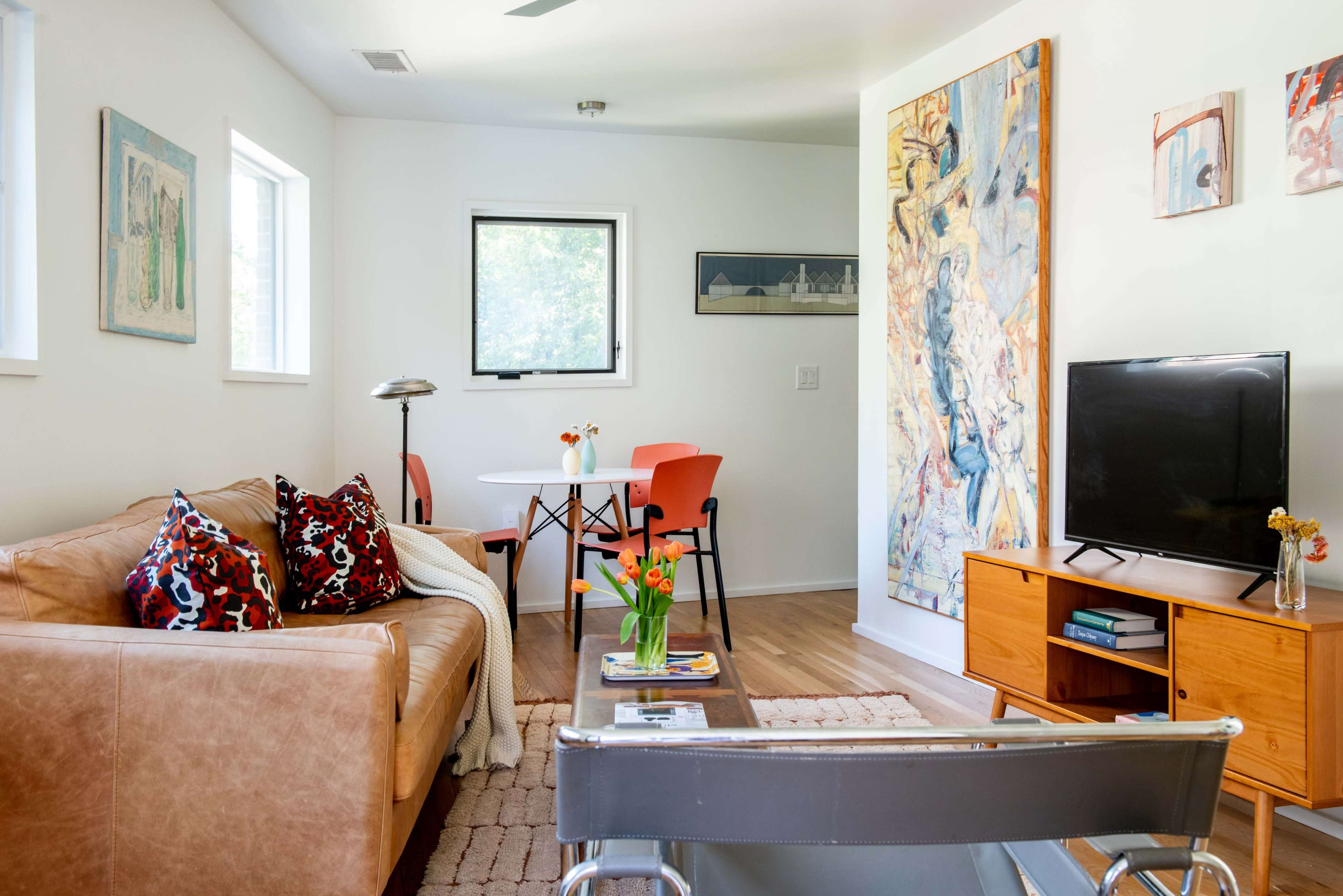 A living room features a brown leather sofa, a wooden coffee table, a flat-screen TV on an orange console, and a dining area with a white table and orange chairs.