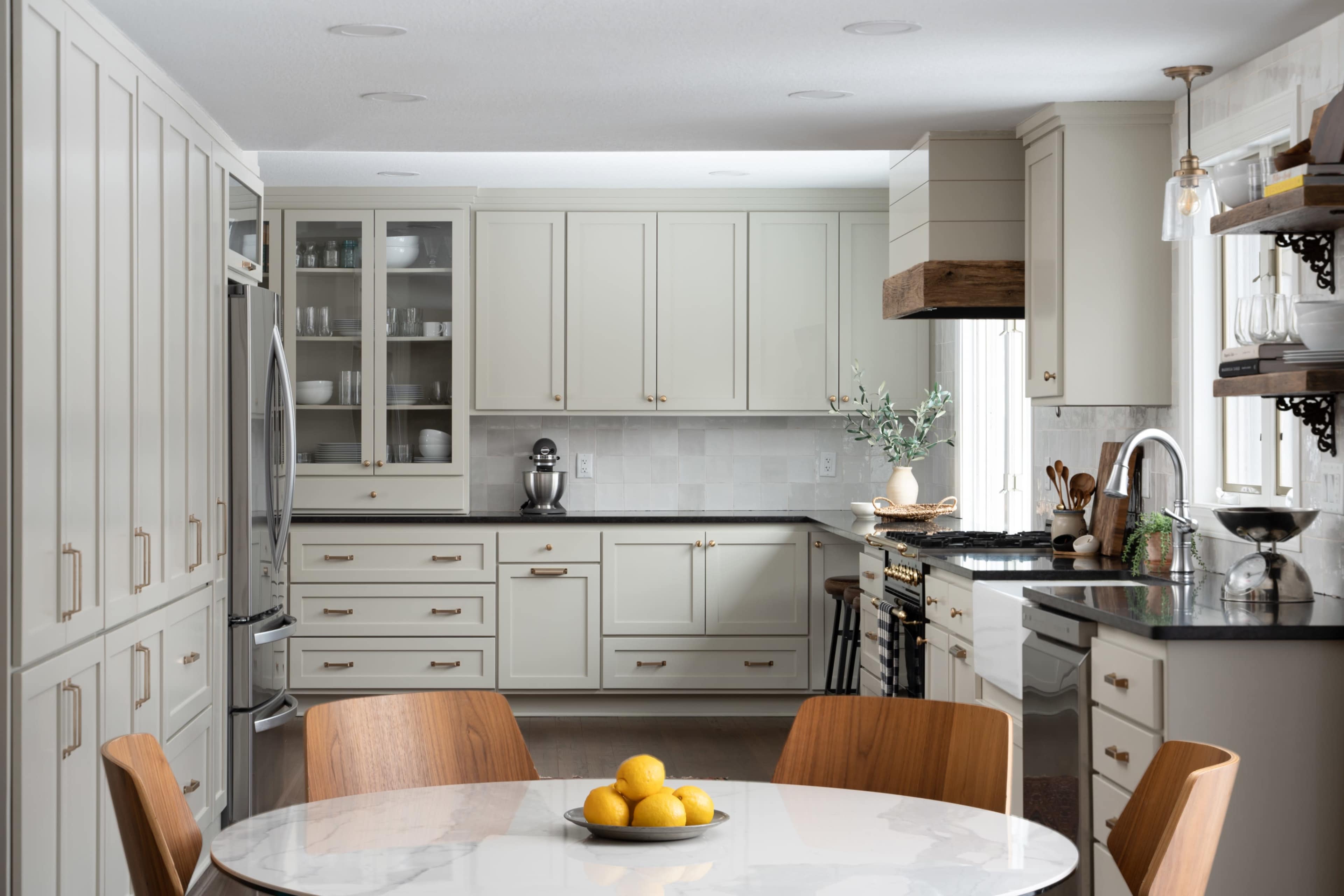 A modern kitchen features light-colored cabinetry, a round dining table with a centerpiece of lemons, and a stainless steel refrigerator.