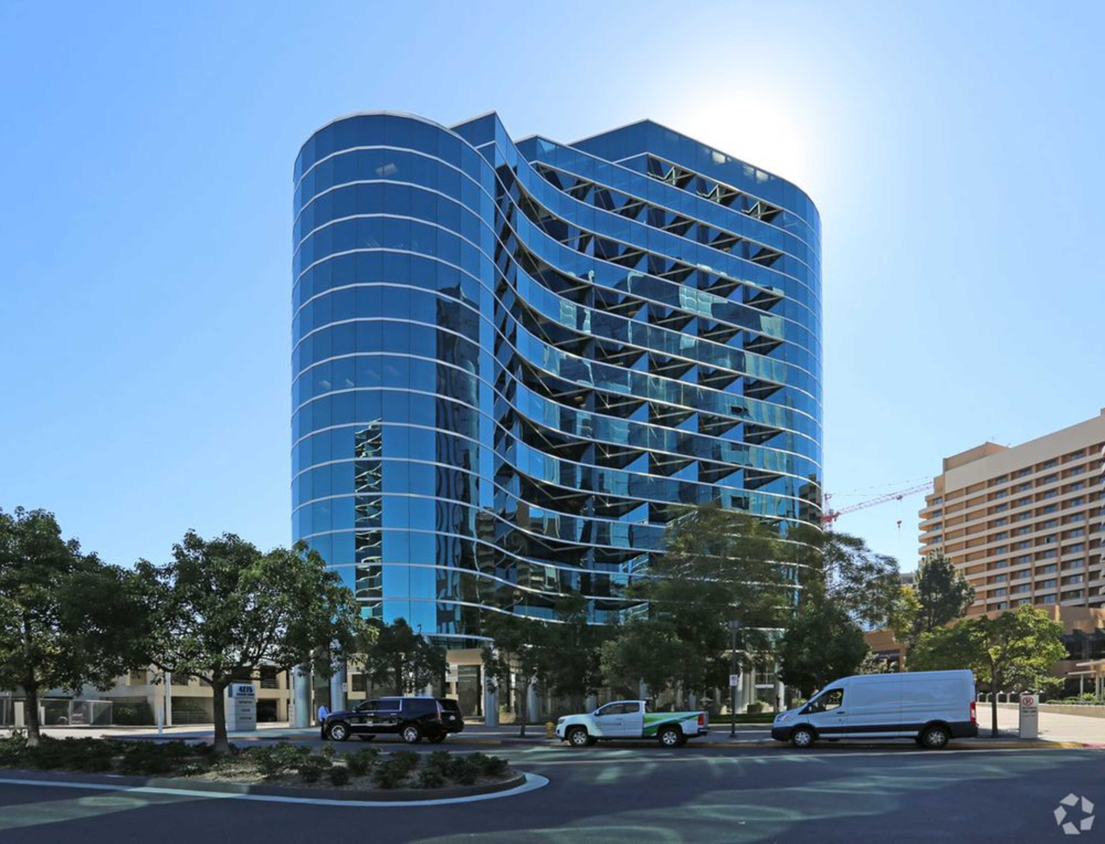 A modern, multi-story glass office building is set against a clear blue sky, reflecting sunlight and surrounded by vehicles and trees.
