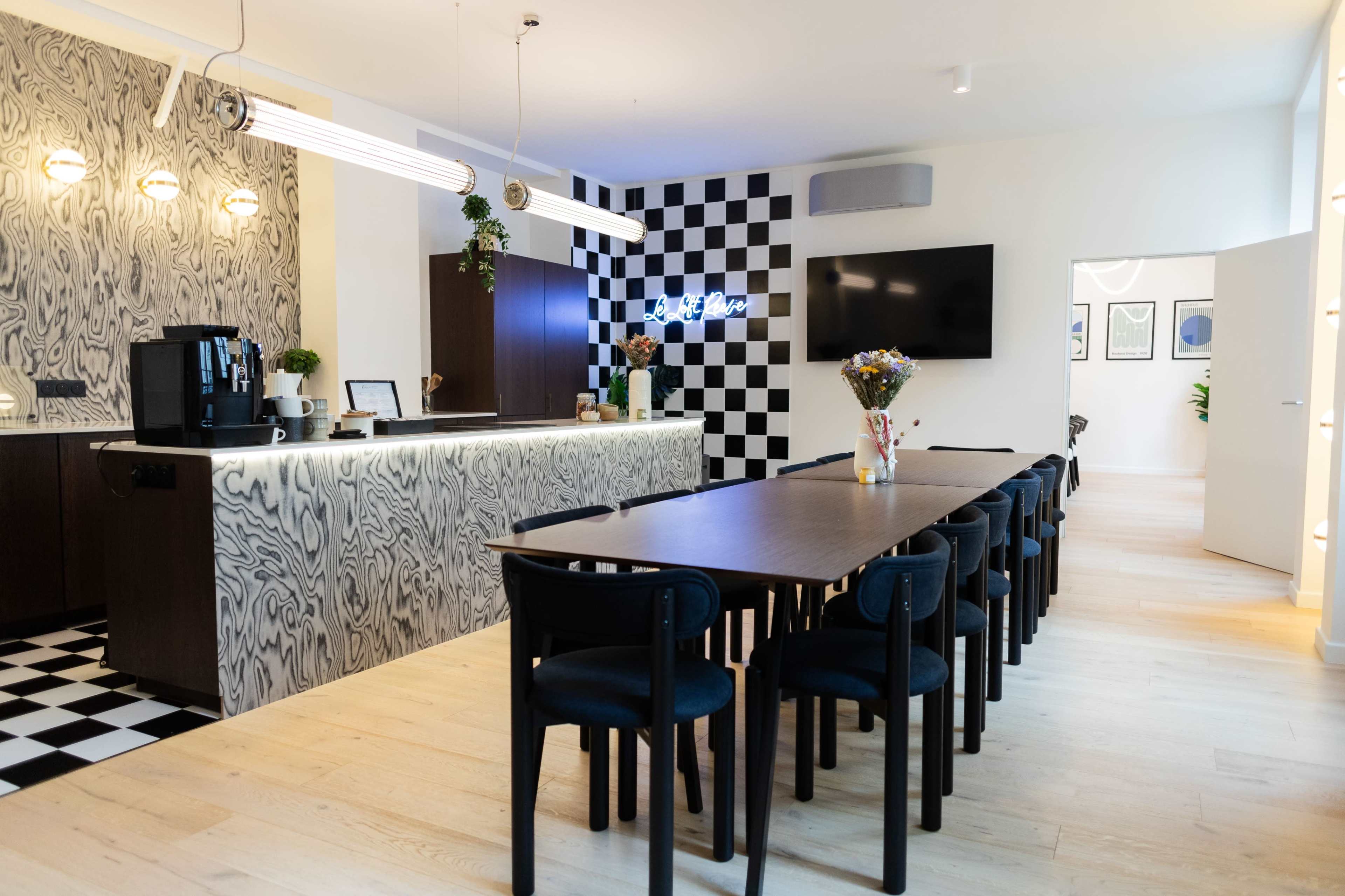 The image shows a modern kitchen area with a large table and black-and-white checkered walls, alongside a coffee station and decorative flowers on the table.