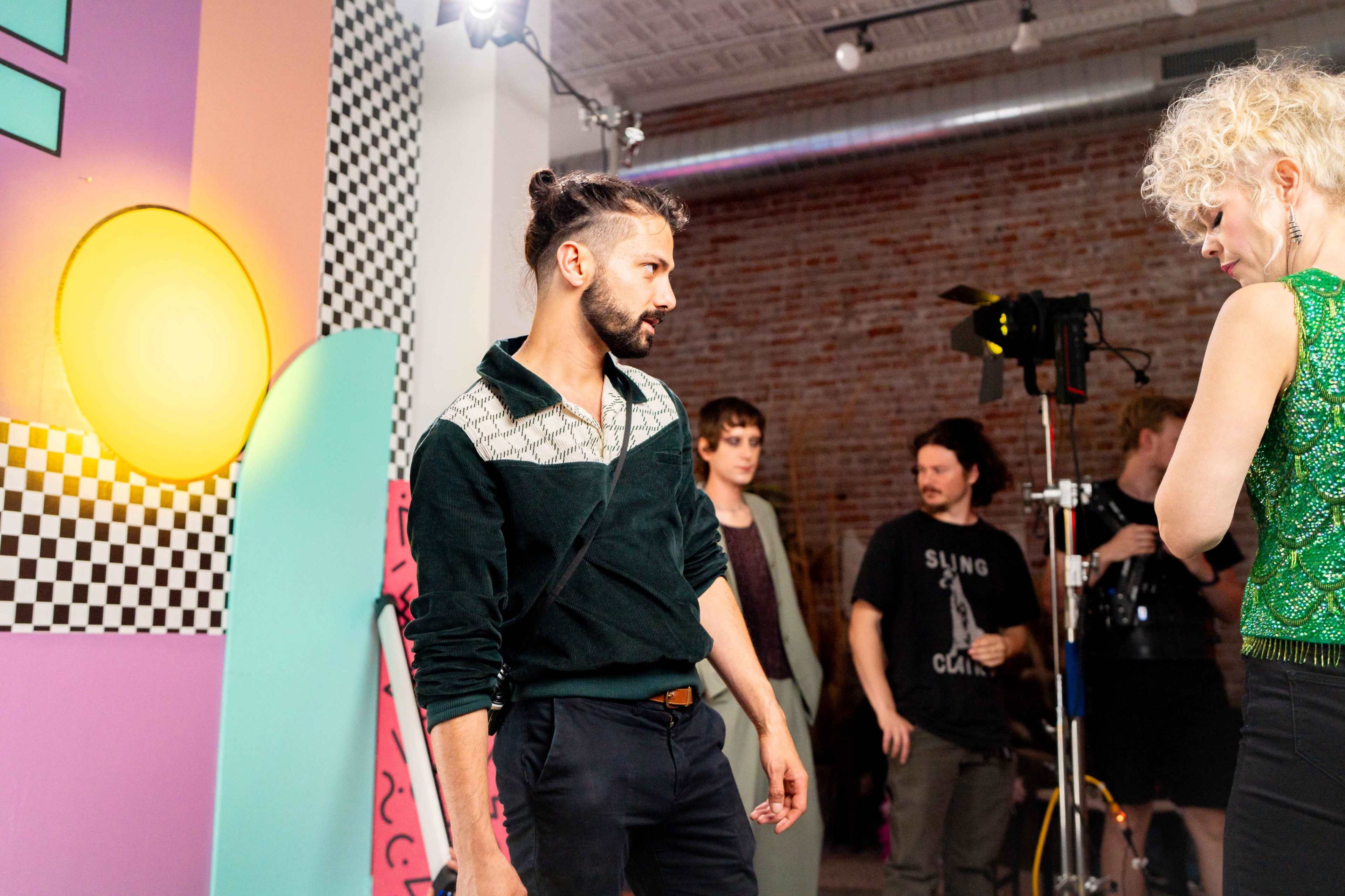 A man in a green zip-up shirt stands in a studio setting with a colorful backdrop, while a woman in a sequined top interacts with him.