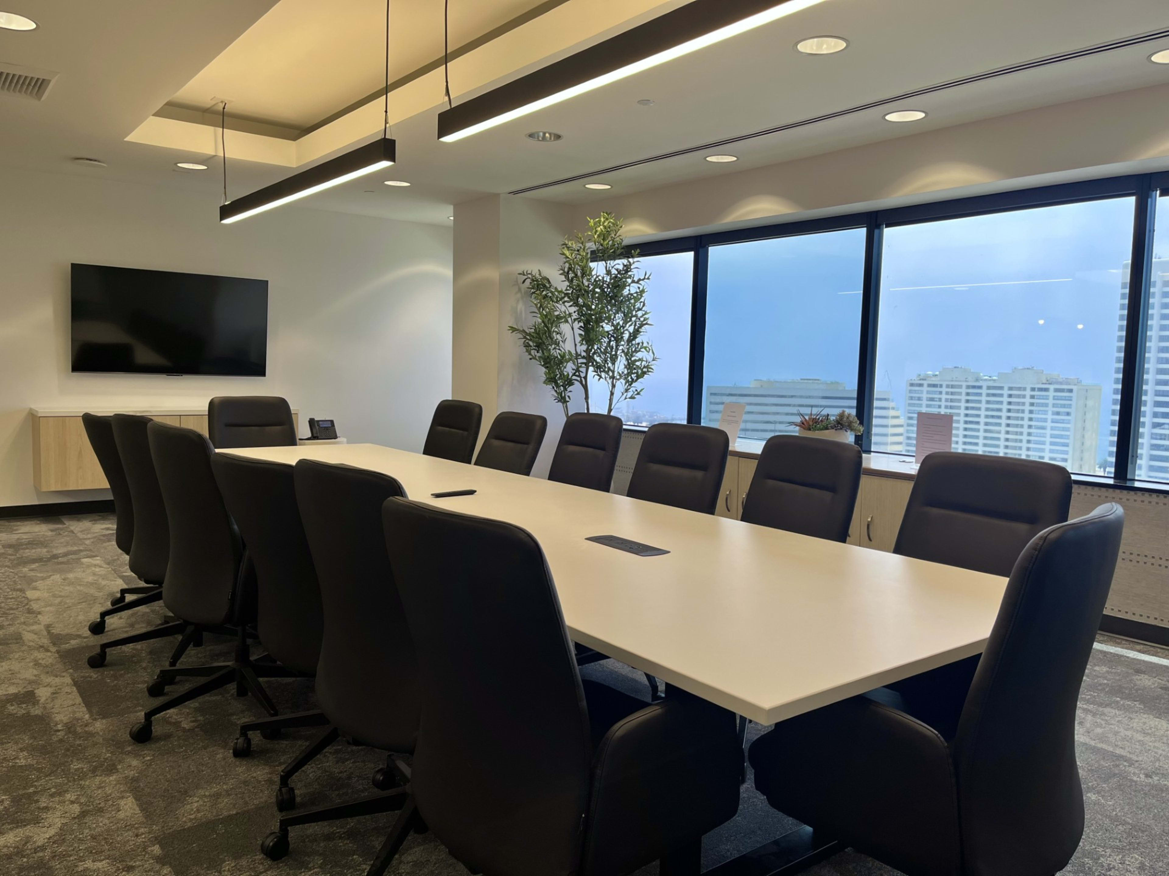 A large conference room features a long white table surrounded by black swivel chairs, with large windows showing a cityscape outside.