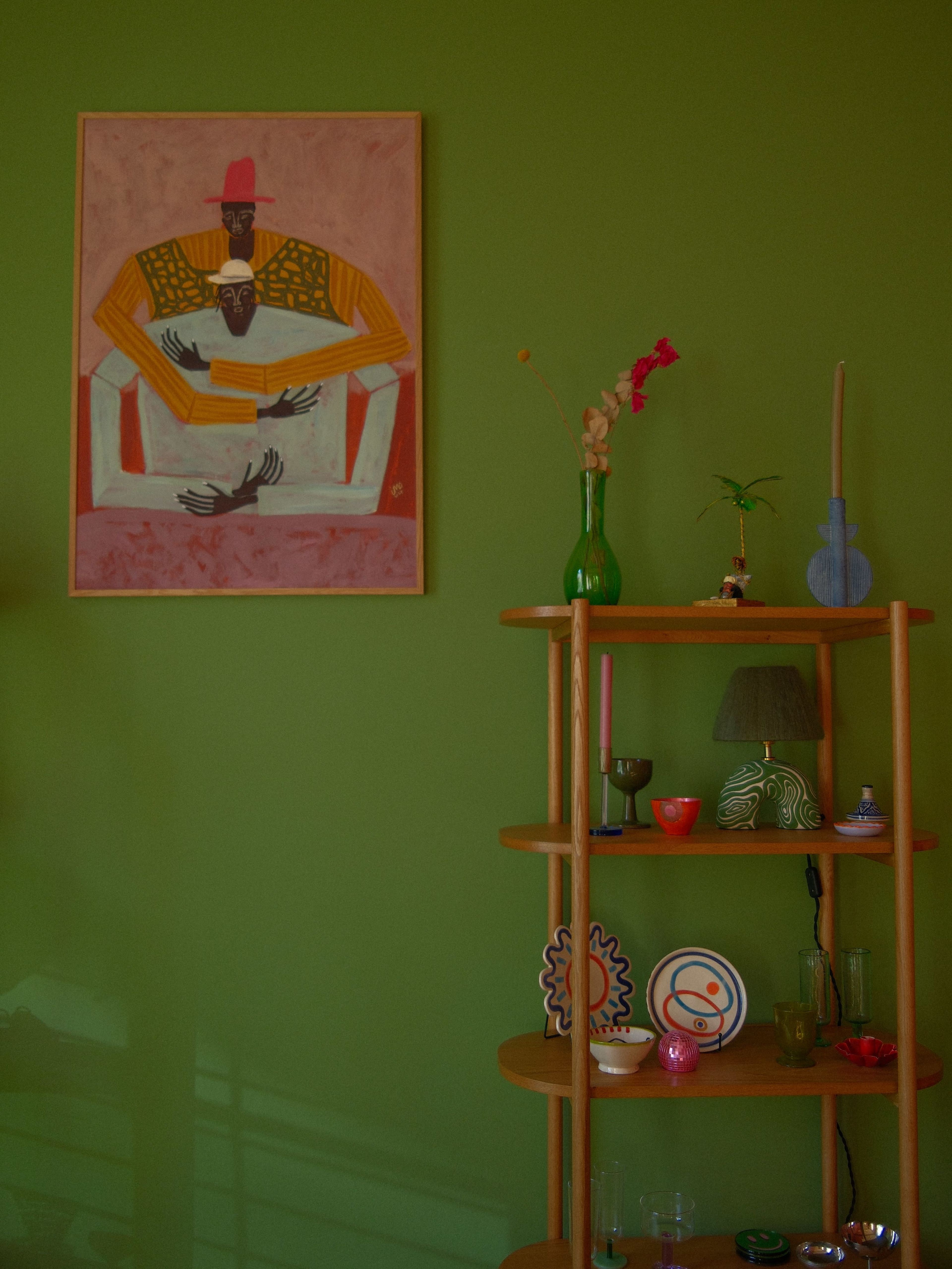 The image features a green wall adorned with a large painting and a wooden shelving unit displaying various decorative items and plants.