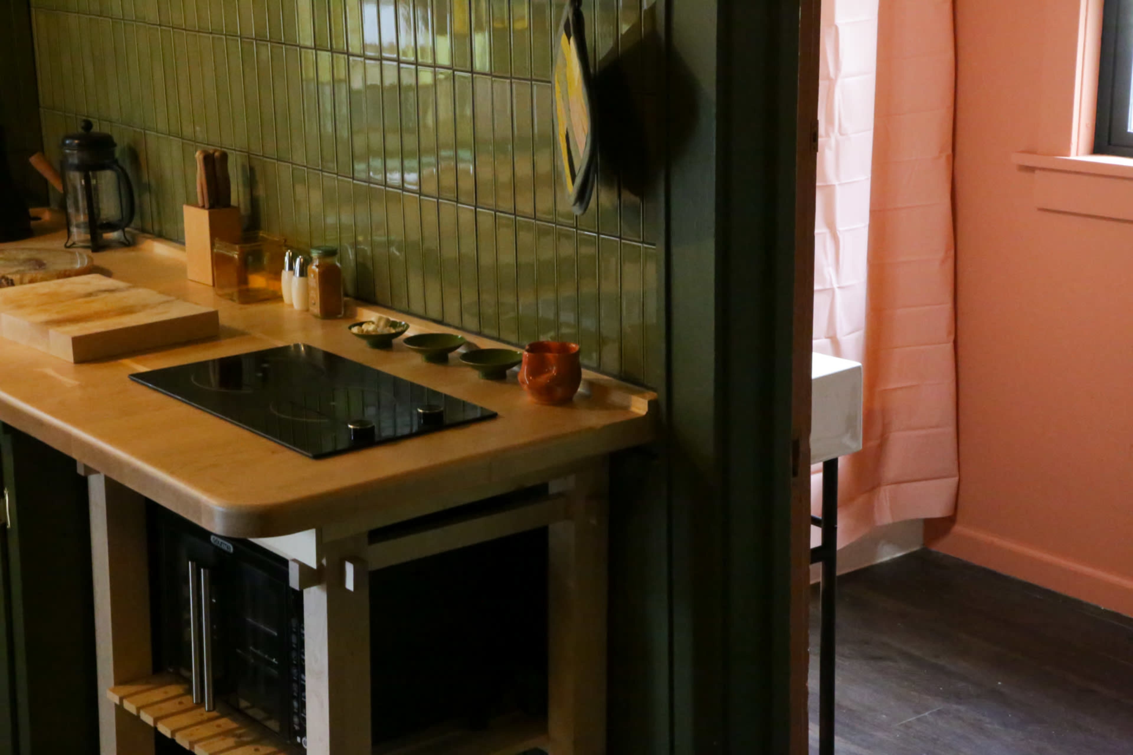 The image shows a modern kitchen with a countertop featuring an induction cooktop, surrounded by green walls and a partially visible pink room in the background.