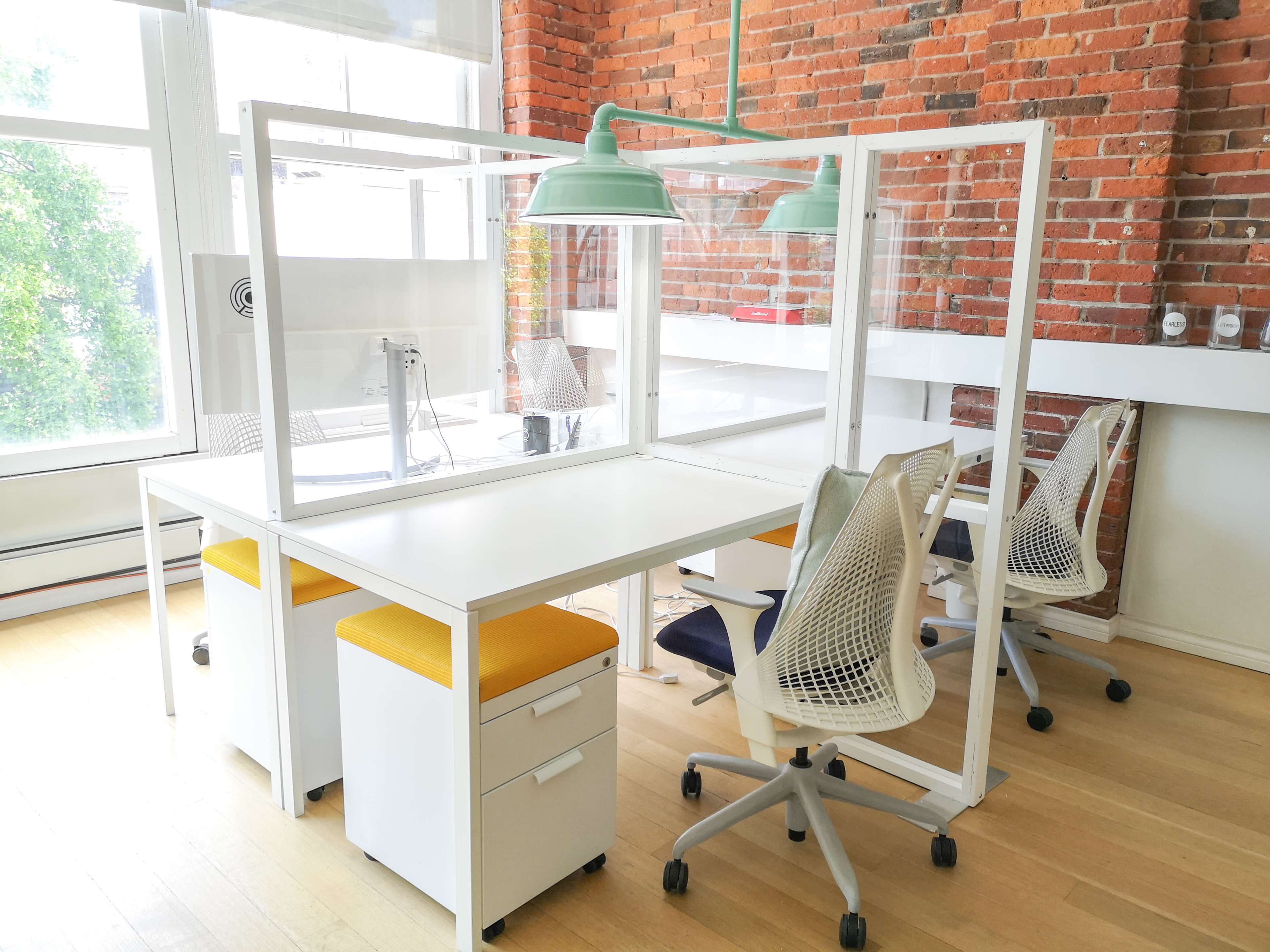 A modern office workspace featuring two desks separated by transparent partitions, with ergonomic chairs and bright yellow seat cushions.