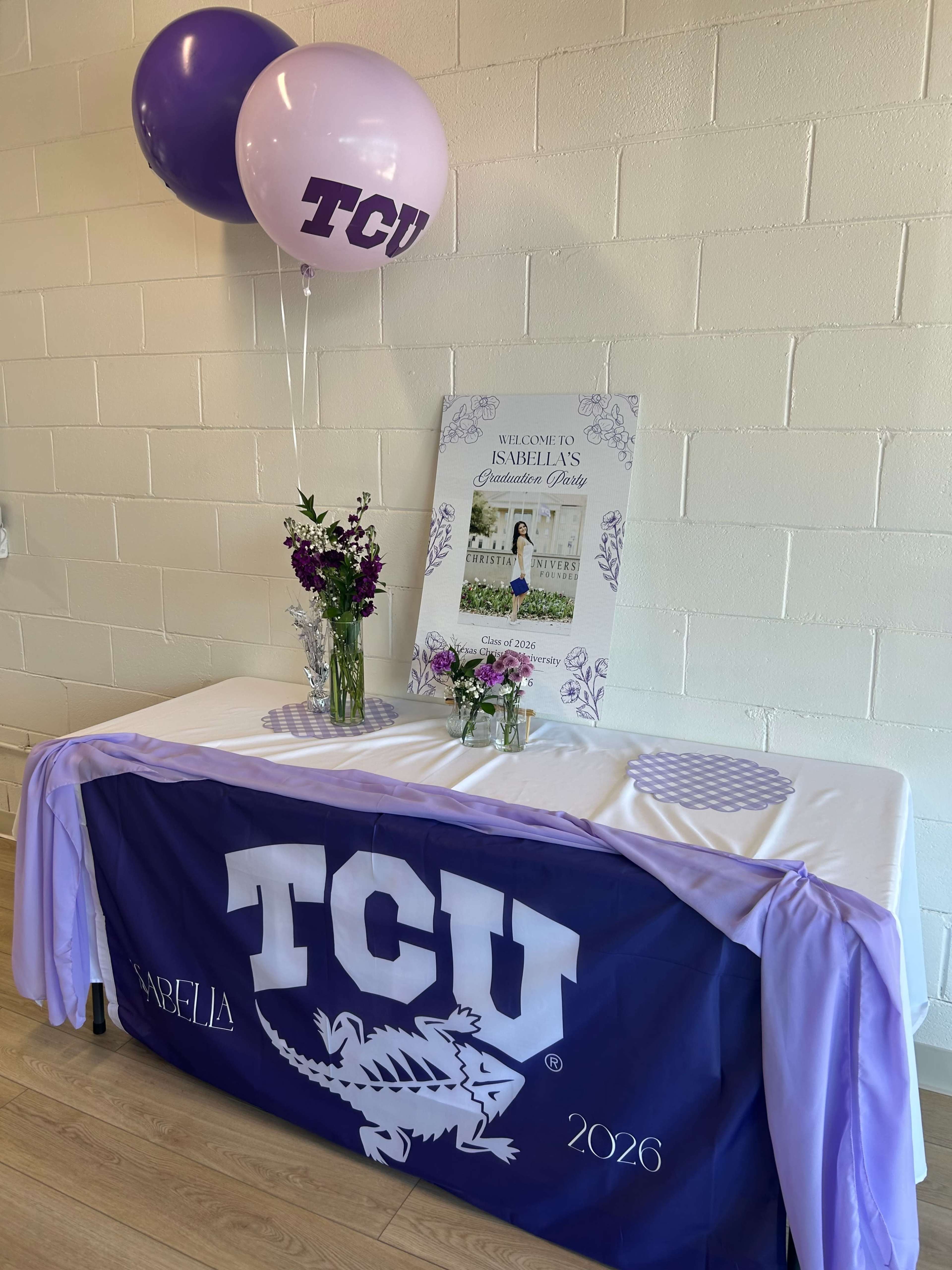 The image shows a decorated table featuring a TCU banner, balloons, a welcome sign, and floral arrangements for a celebration.