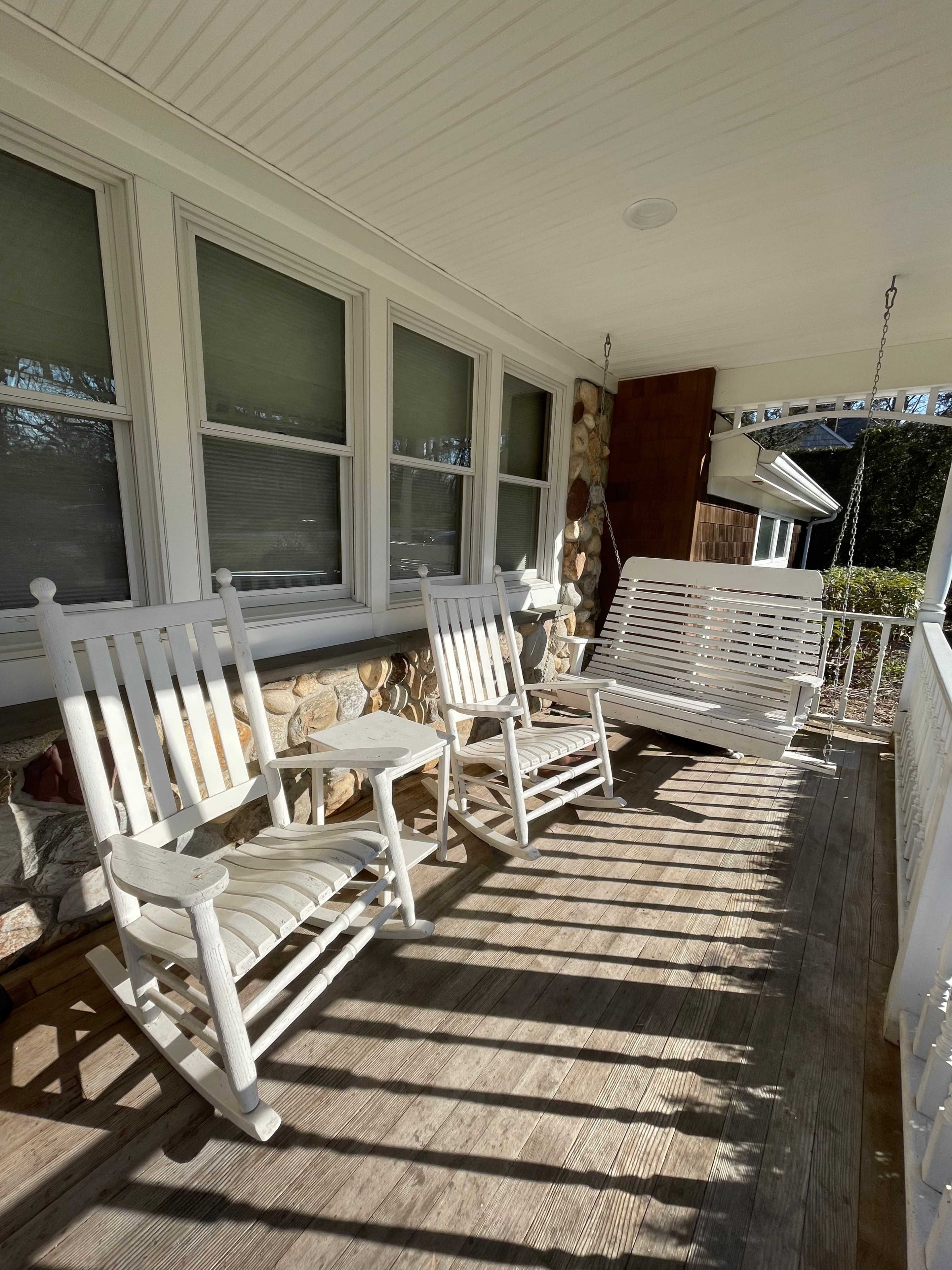 A porch with two white rocking chairs and a white swing, positioned on a wooden floor and surrounded by stone walls.