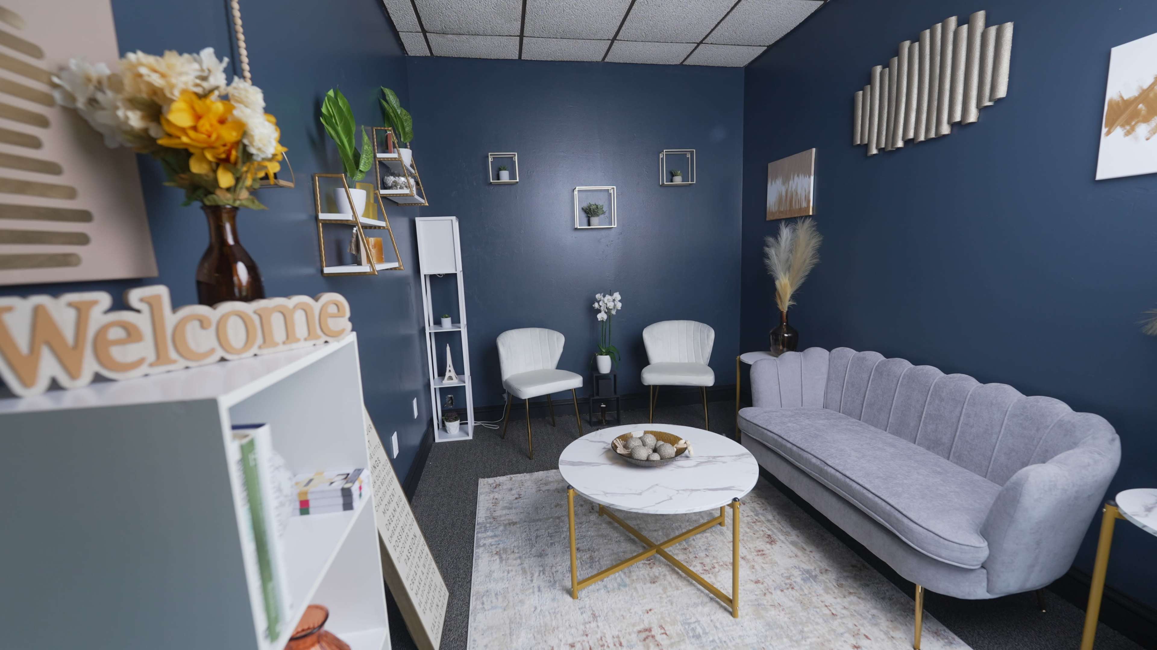 The image shows a cozy waiting area featuring a gray couch, two white chairs, a round coffee table, and decorative accents against a dark blue wall.