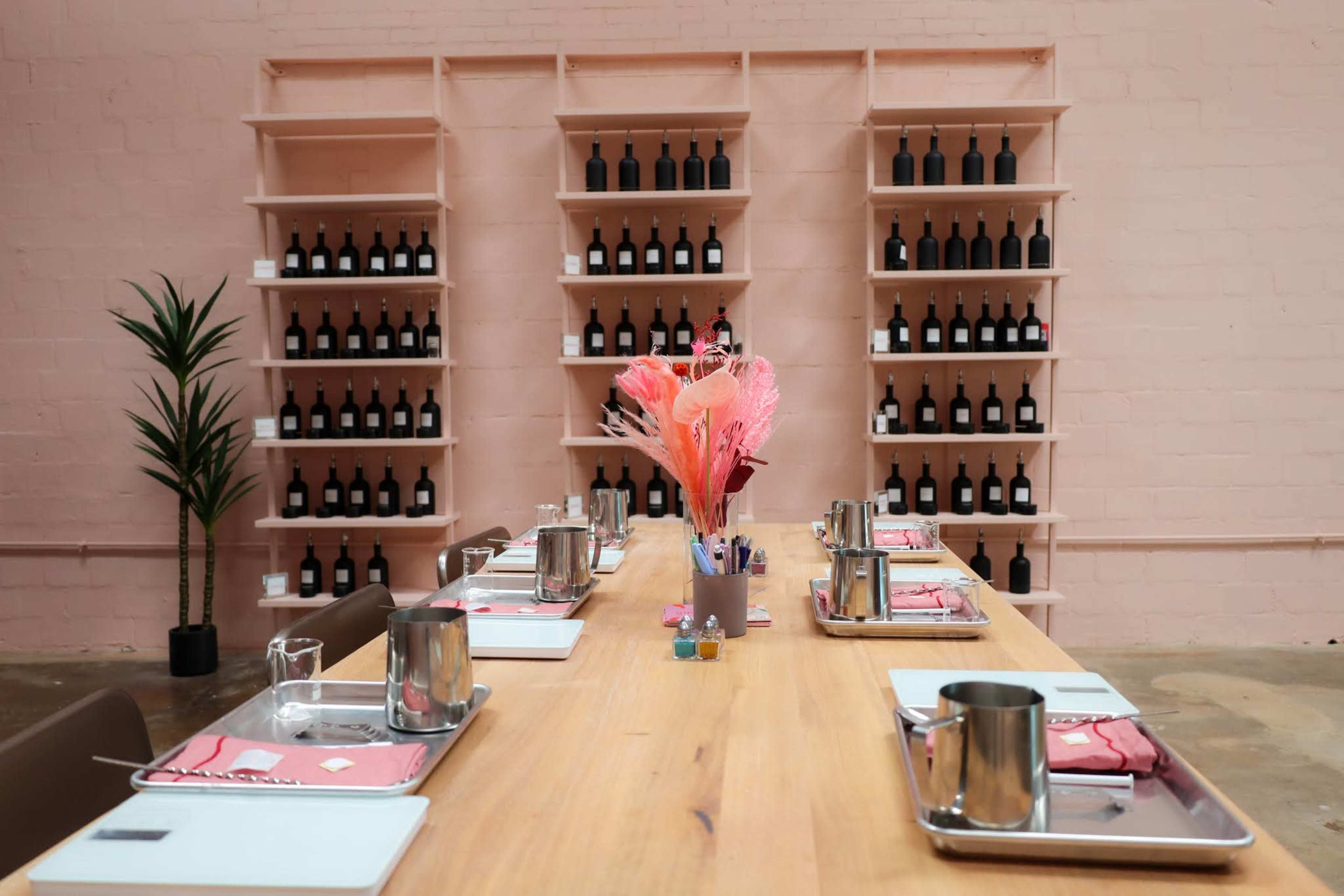 A long wooden table is set with metal trays and napkins, facing shelves filled with wine bottles against a pink wall.