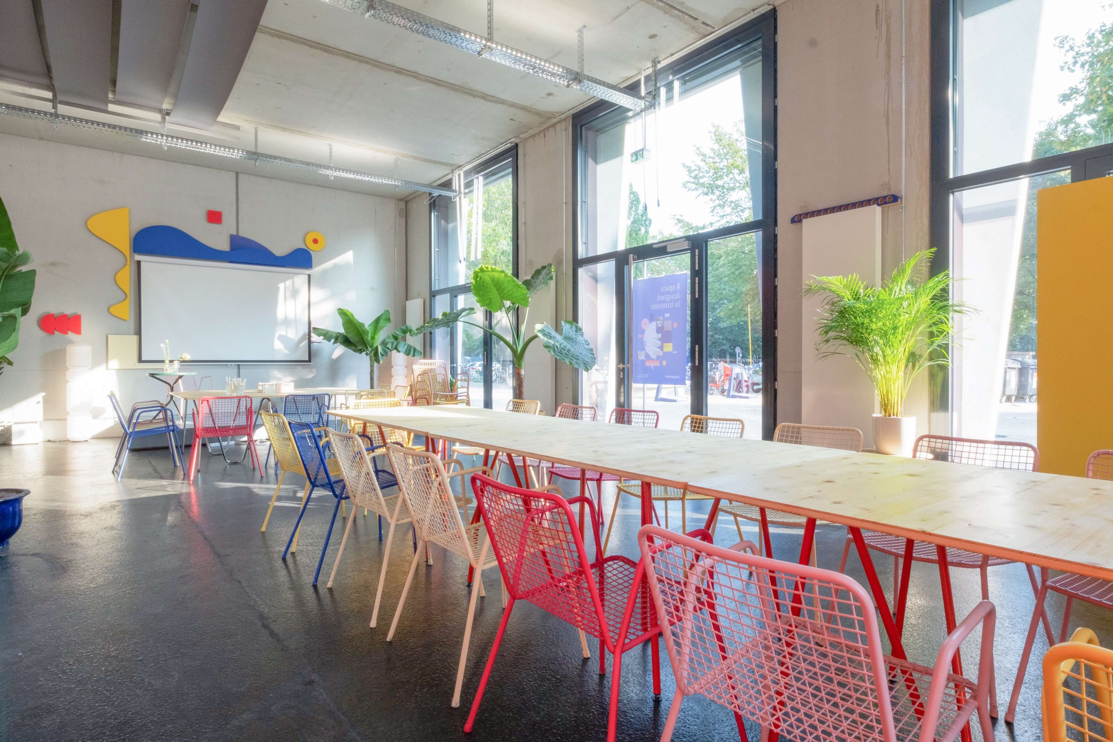 A long table with colorful chairs is set in a well-lit room featuring large windows and plants, with a projector screen at one end.