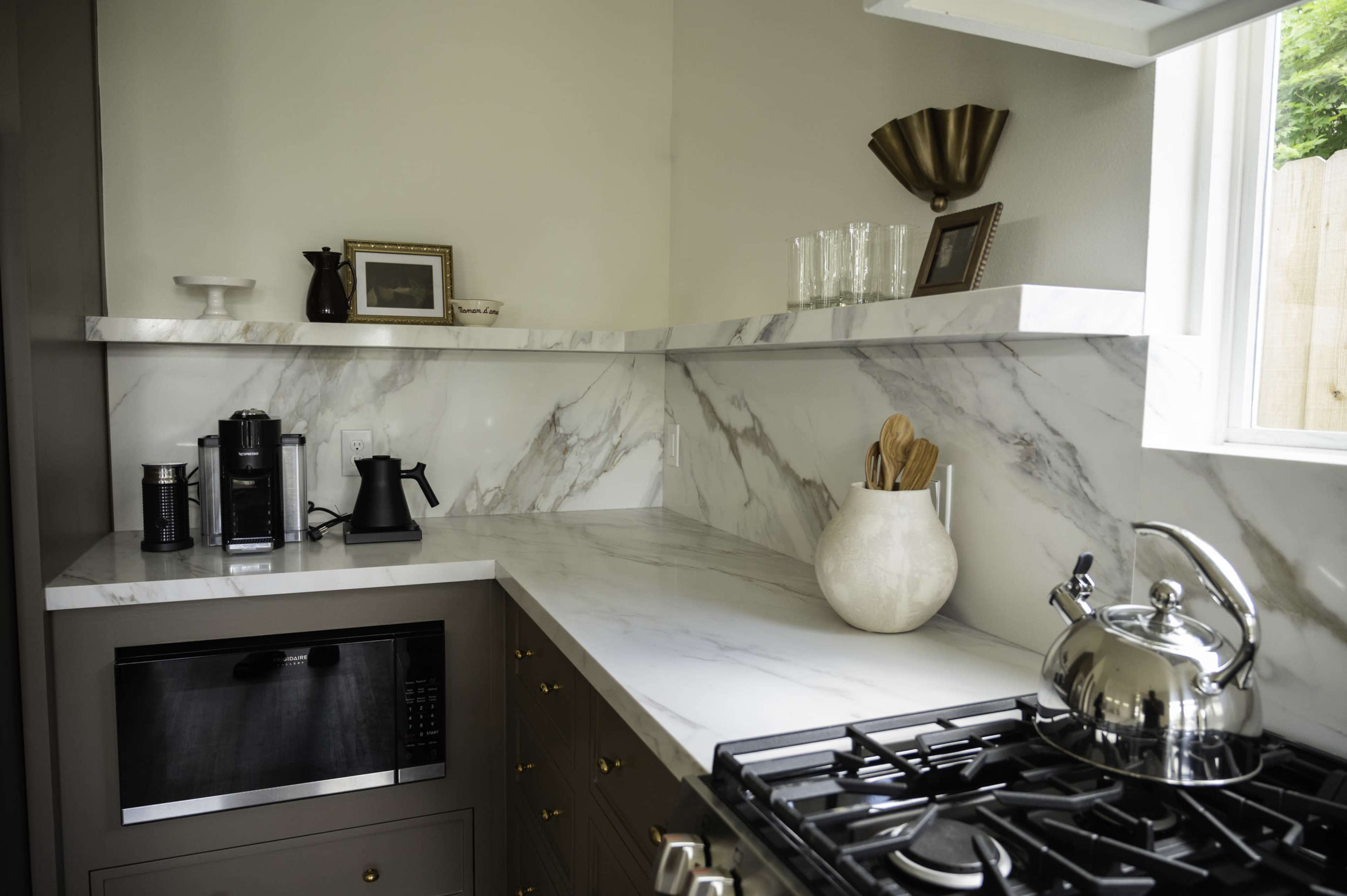 The image shows a modern kitchen with marble countertops, a microwave, a kettle, and kitchen utensils displayed on open shelves.